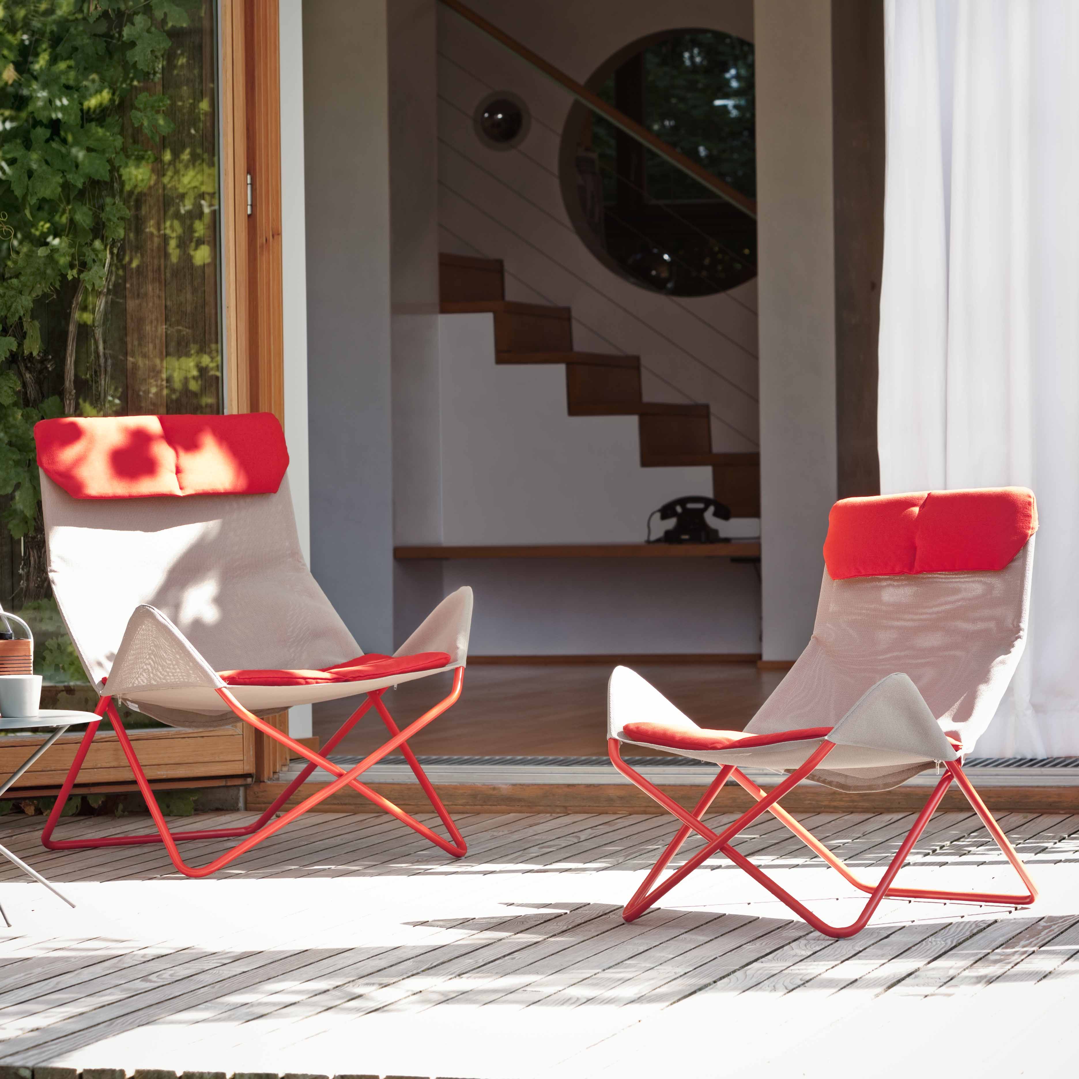 Two In-Out Chairs by Richard Lampert, featuring modern, weather-resistant designs with red frames and headrests, sit on a wooden deck near a sliding door. Inside the house, you can spot a staircase and retro phone. Ideal for outdoor use year-round.