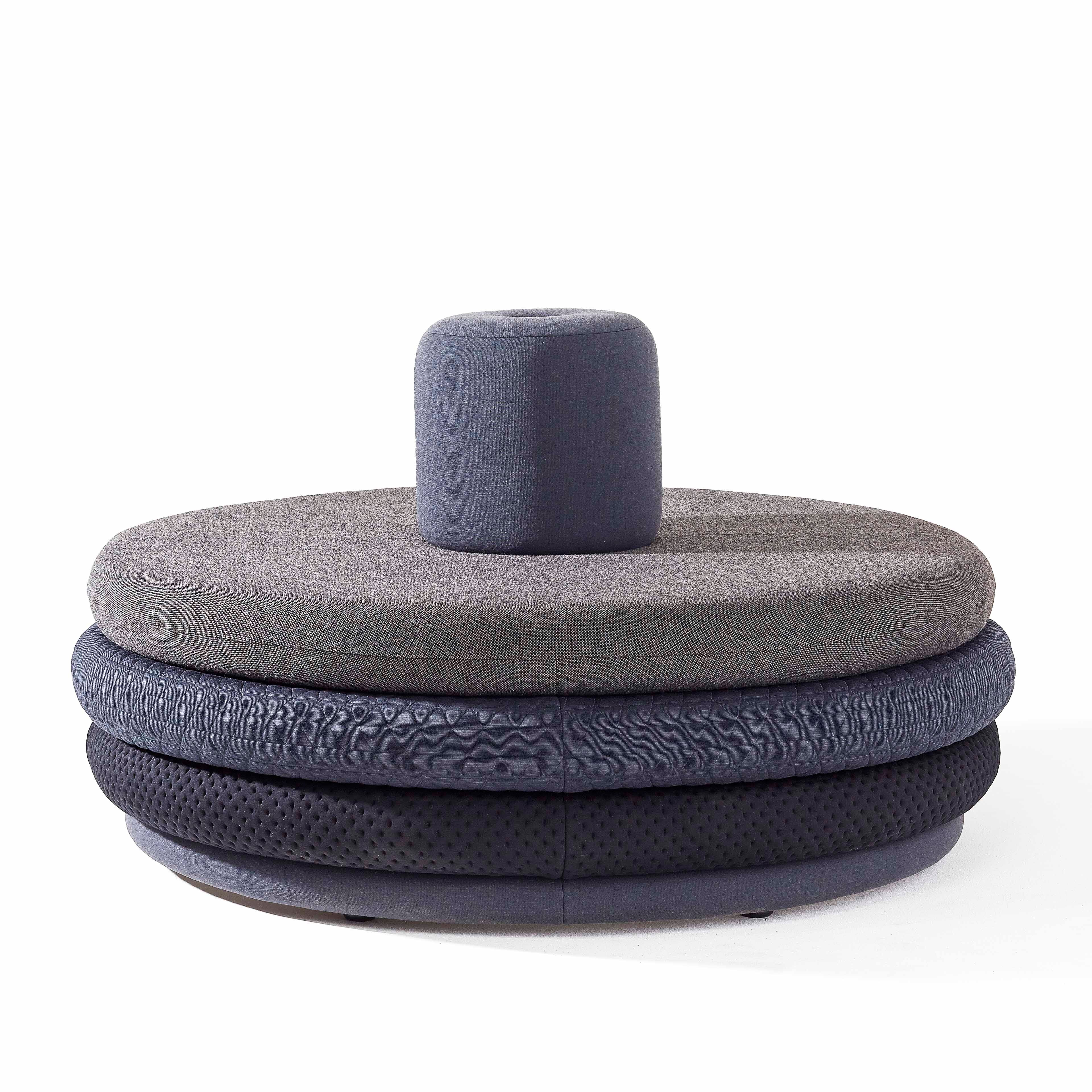 Richard Lampert presents the Island Tom, a modern circular ottoman with layered textured upholstery in gray and black. Reminiscent of personalized stools, it features a dark gray cylindrical cushion against a white background.
