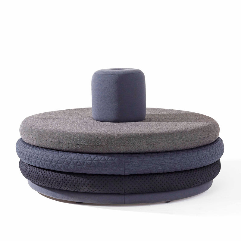 Richard Lampert presents the Island Tom, a modern circular ottoman with layered textured upholstery in gray and black. Reminiscent of personalized stools, it features a dark gray cylindrical cushion against a white background.