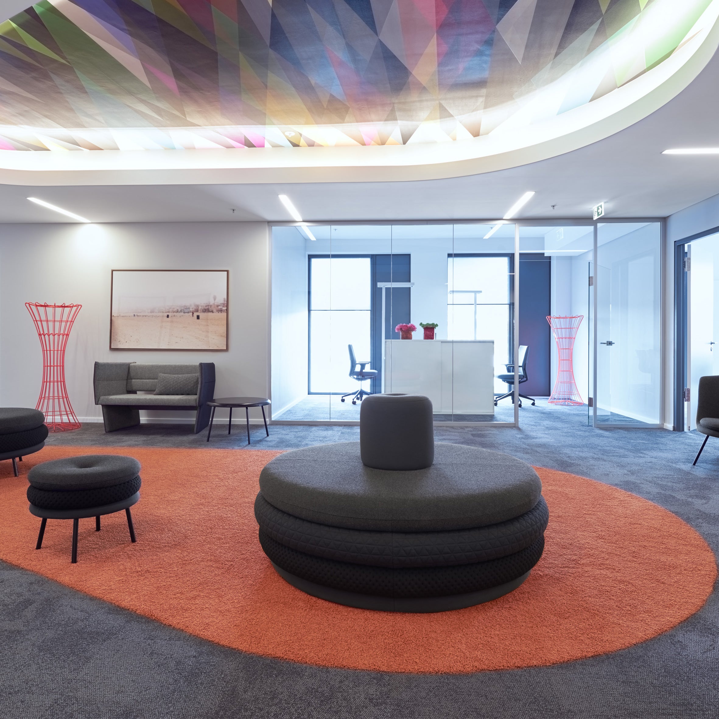 Modern office space with a vibrant geometric ceiling and glass-walled office featuring Richard Lamperts Island Tom minimalistic furniture, personalized stools, a large orange rug, gray poufs and couch, red flower pots on a white desk, and abstract wall art.