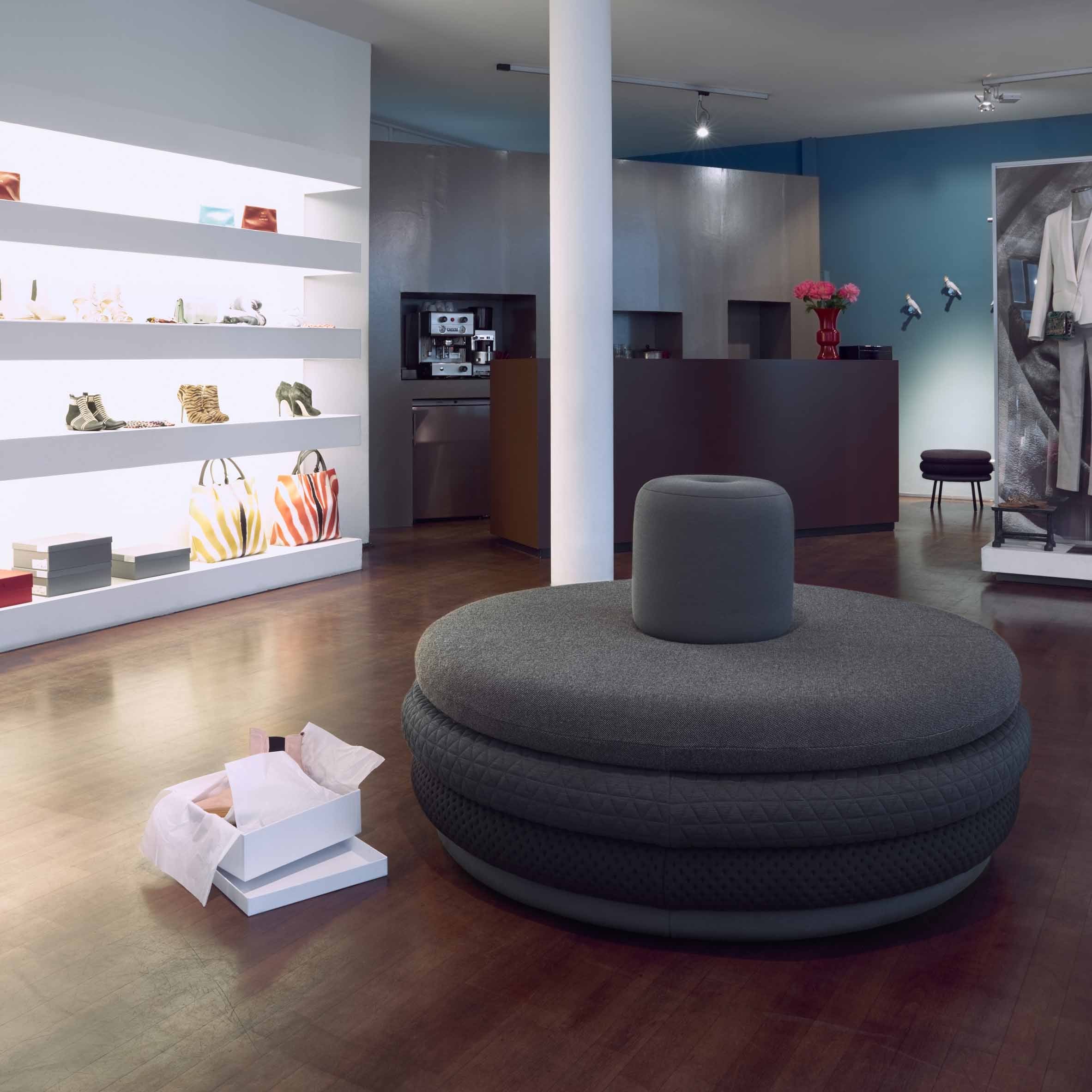 A modern boutique interior showcases a gray round seating area with Richard Lamperts Island Tom stools. Shelves of bags and shoes line the left wall, a counter with pink flowers is at the back, clothes hang on the right wall, and wooden flooring enhances the decor.