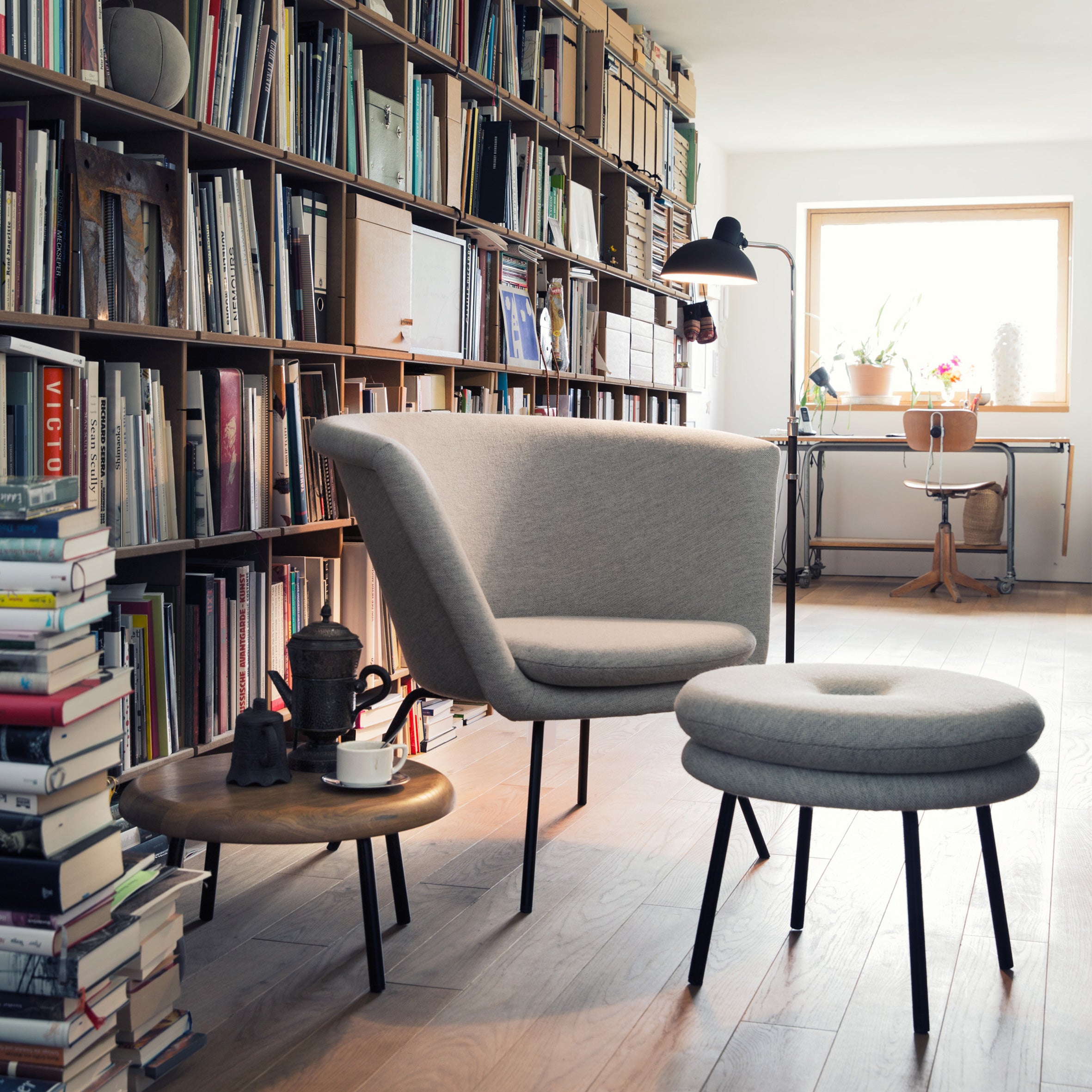A cozy reading nook features a grey armchair with a matching ottoman beside a Richard Lampert Little Tom Stool. Bookshelves packed with books frame the scene, while a small round table with an oak top supports a coffee cup. A window floods natural light over the desk in the background.
