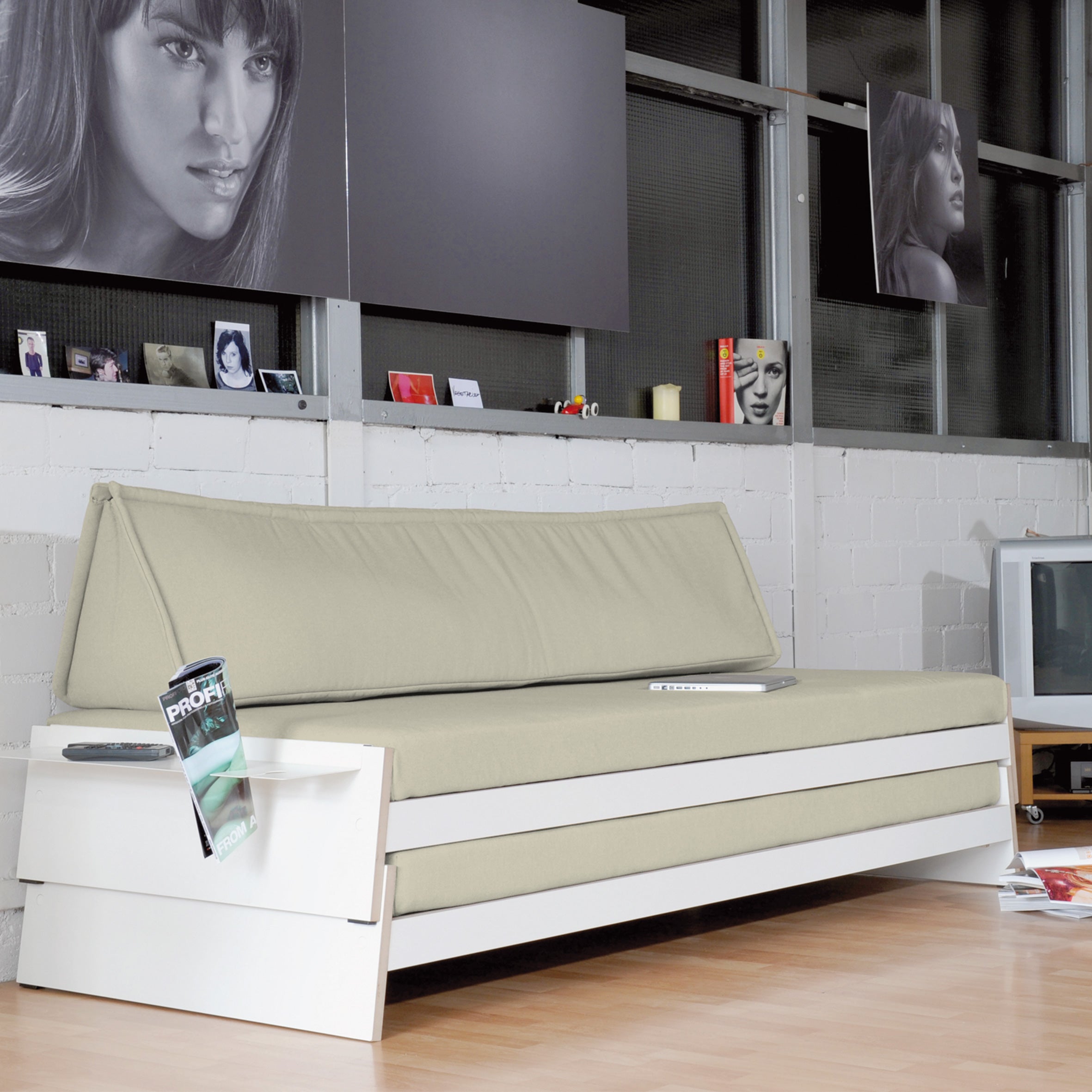 A modern room features a beige Lönneberga Stacking Bed by Richard Lampert and a versatile white side table adorned with photos and a TV. Large windows highlight the brick wall, while framed black-and-white portraits add elegance, with a magazine on the table enhancing its chic charm.