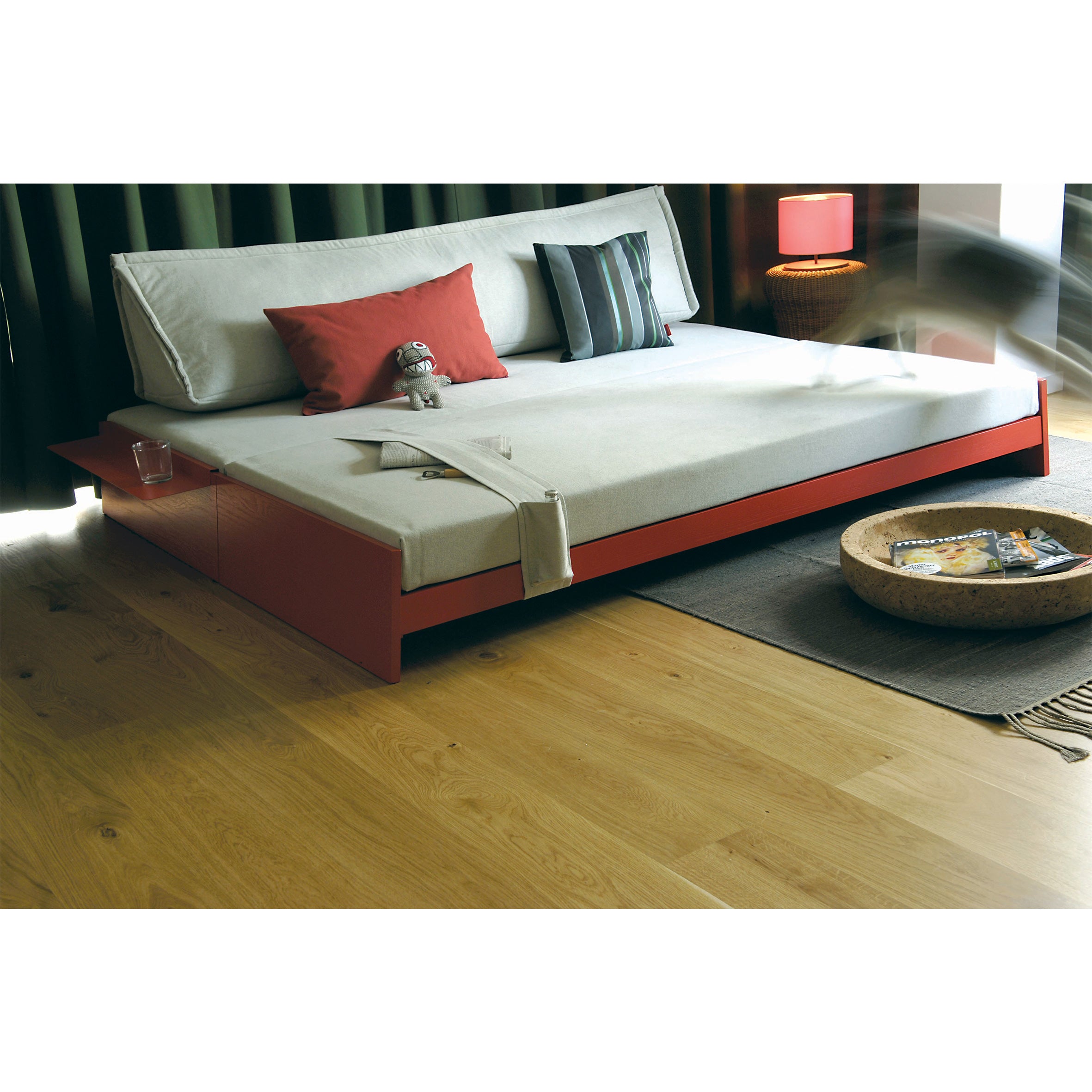 A modern bedroom showcases space efficiency with the Lönneberga Stacking Bed by Richard Lampert, adorned in white and red bedding with decorative pillows. A small red lamp on a side table, wooden flooring, and a rug holding a magazine basket complete this cozy space.