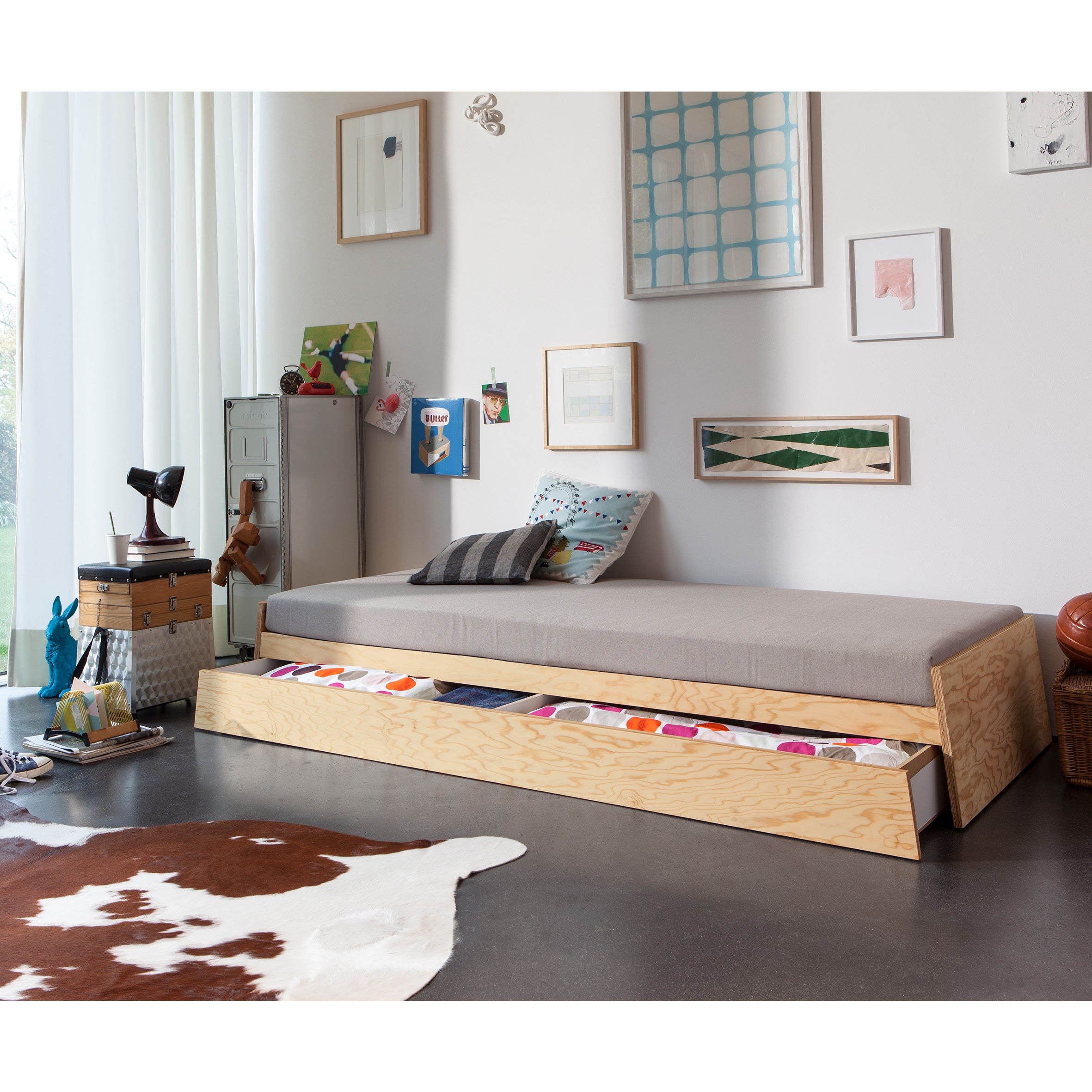A modern bedroom features the versatile Lönneberga Stacking Bed by Richard Lampert, complete with a pull-out multicolored drawer. The space is decorated with artwork, eclectic items like books and a vintage fan, and includes a plush cowhide rug, all illuminated by light streaming through a large window.