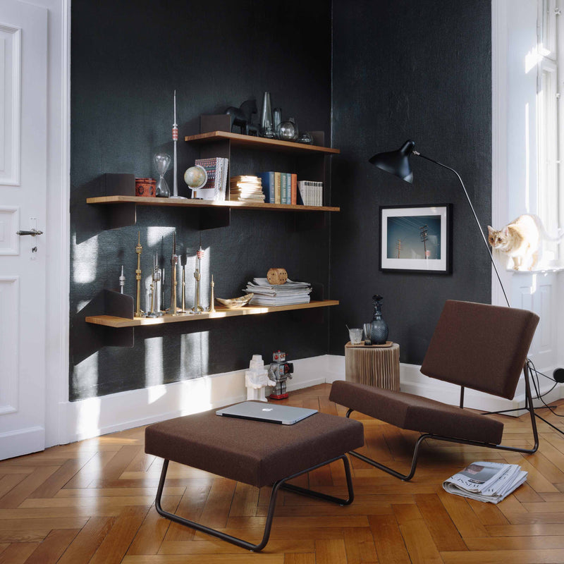 A cozy room with dark walls and wooden floors features a Hirche Lounge Ottoman by Richard Lampert, wall-mounted shelves with books and decor, a modern floor lamp, and a cat on the windowsill. Sunlight streams in, enhancing the warm atmosphere of this Bauhaus-inspired space.