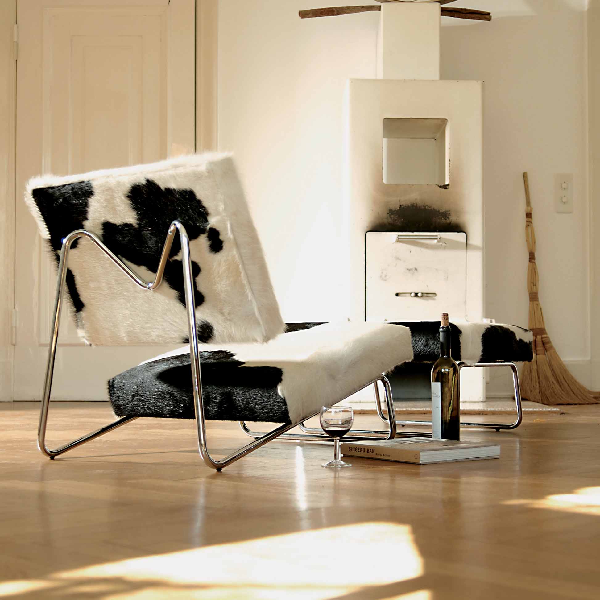 A modern living room showcases the Hirche Lounge Ottoman by Richard Lampert, featuring black and white fur upholstery on a sleek metal frame, reminiscent of Bauhaus design. On the floor are a wine bottle, glass, and book. A broom leans against the wall beside a small white cabinet.