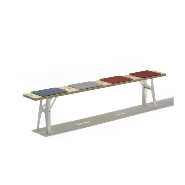 The Ludwig Bench by Richard Lampert is a sustainable long wooden bench with white metal legs, featuring four square padded seats in blue, gray, and two shades of red. Reminiscent of a beer tables sturdy design, its ideal for outdoor settings against a plain white backdrop.