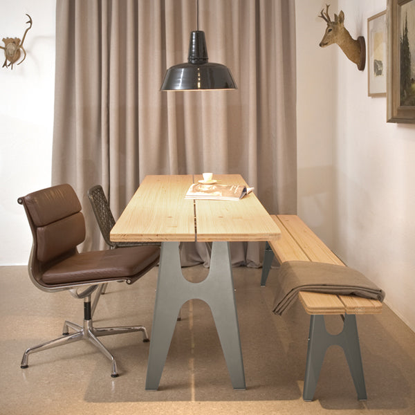 A modern dining area showcases the Richard Lampert Ludwig Table with foldable legs, a bench, and a brown leather swivel chair beneath a black pendant light. Beige curtains adorn the wall, while a deer head mount and framed artwork complete the decor.