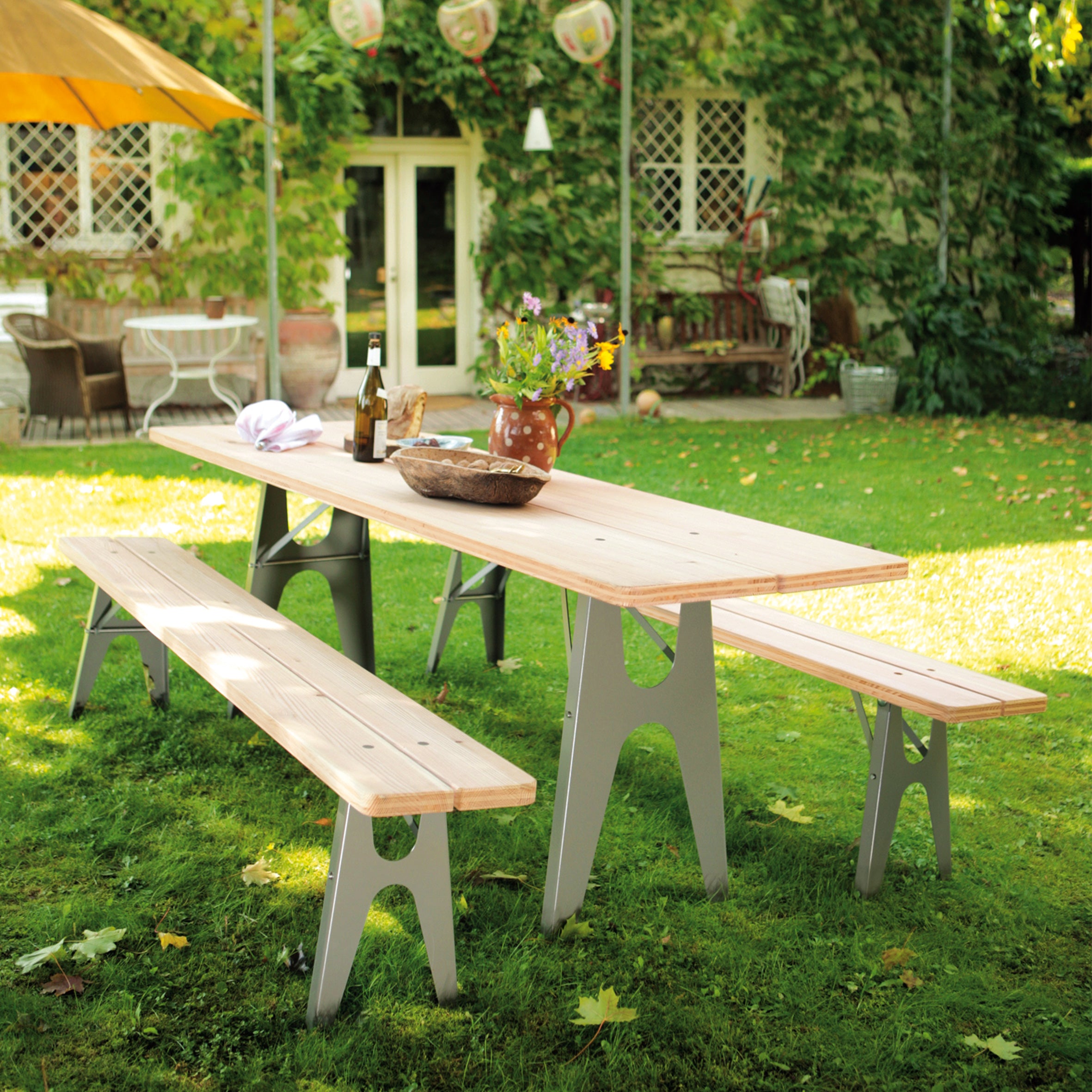 In a sunlit garden, the Richard Lampert Ludwig Table with weather-resistant foldable legs stands ready for festivity. A jug of flowers, wine bottle, and cheese board invite guests. Greenery frames the scene, while an umbrella peeks in from the left.