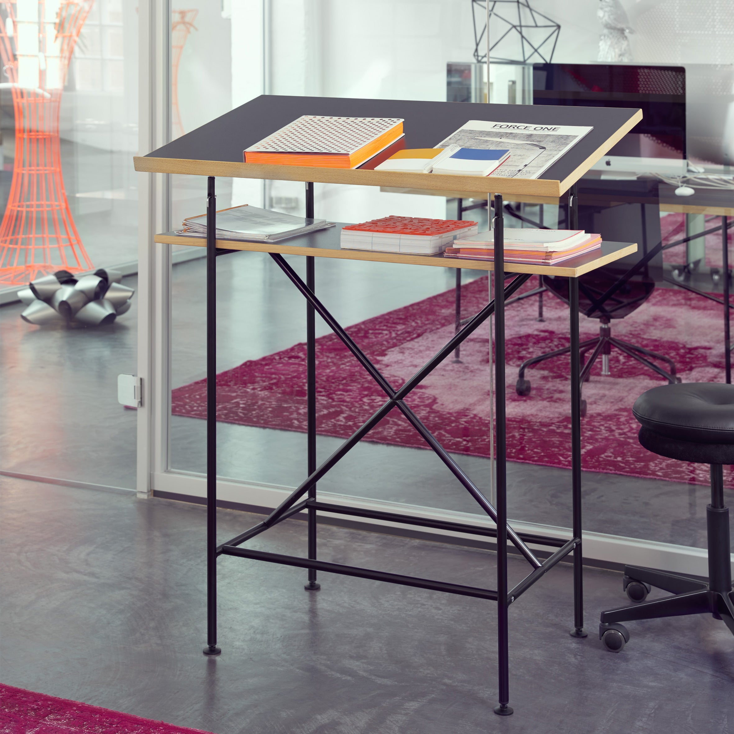 The Milla High Desk by Richard Lampert is a modern Eiermann table featuring a slanted black work surface and lower shelf, set in a bright office with red carpet and glass walls. Paired with a black chair, it’s ideal for active sitting and standing.
