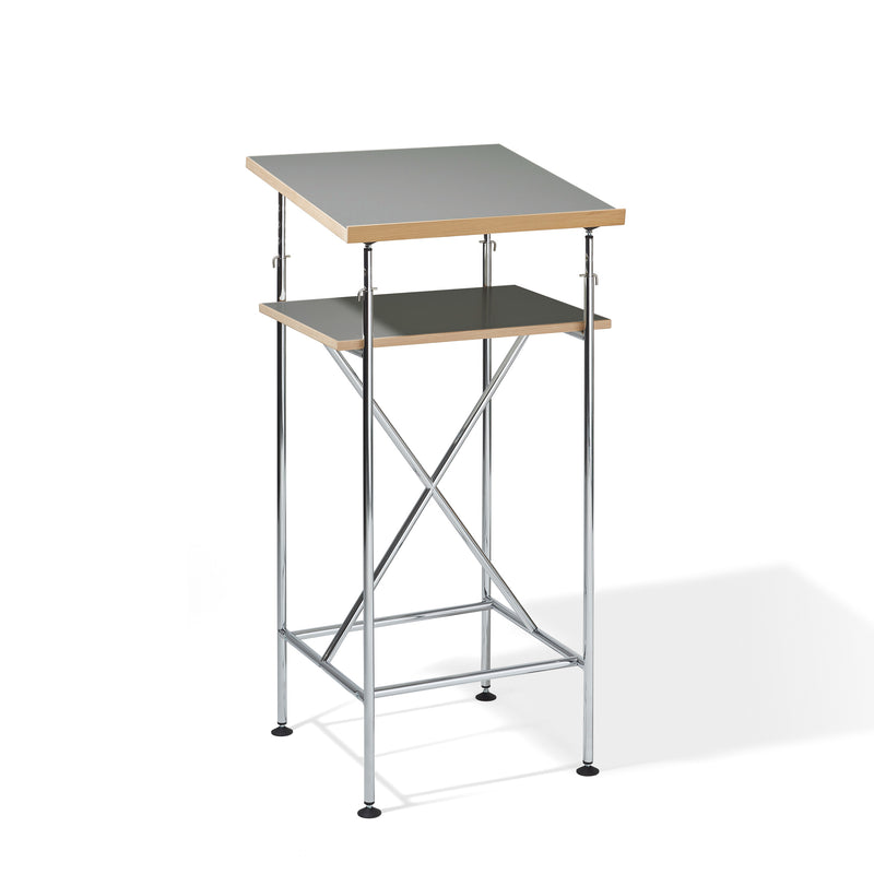 The Milla High Desk by Richard Lampert is a two-tier drafting table inspired by the Eiermann design, featuring a metal frame with adjustable gray tabletops. The top surface angles slightly, while the lower shelf stays flat. It includes stability-enhancing crossbars and legs finished with black feet.