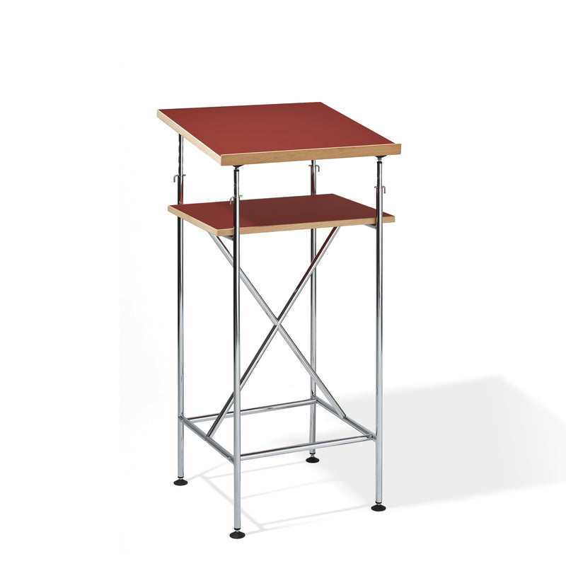 The Milla High Desk by Richard Lampert features a sleek, minimalist design inspired by the Eiermann table, with a red tabletop, wooden lower shelf, and sturdy chrome metal frame with crossbars for active sitting or standing.