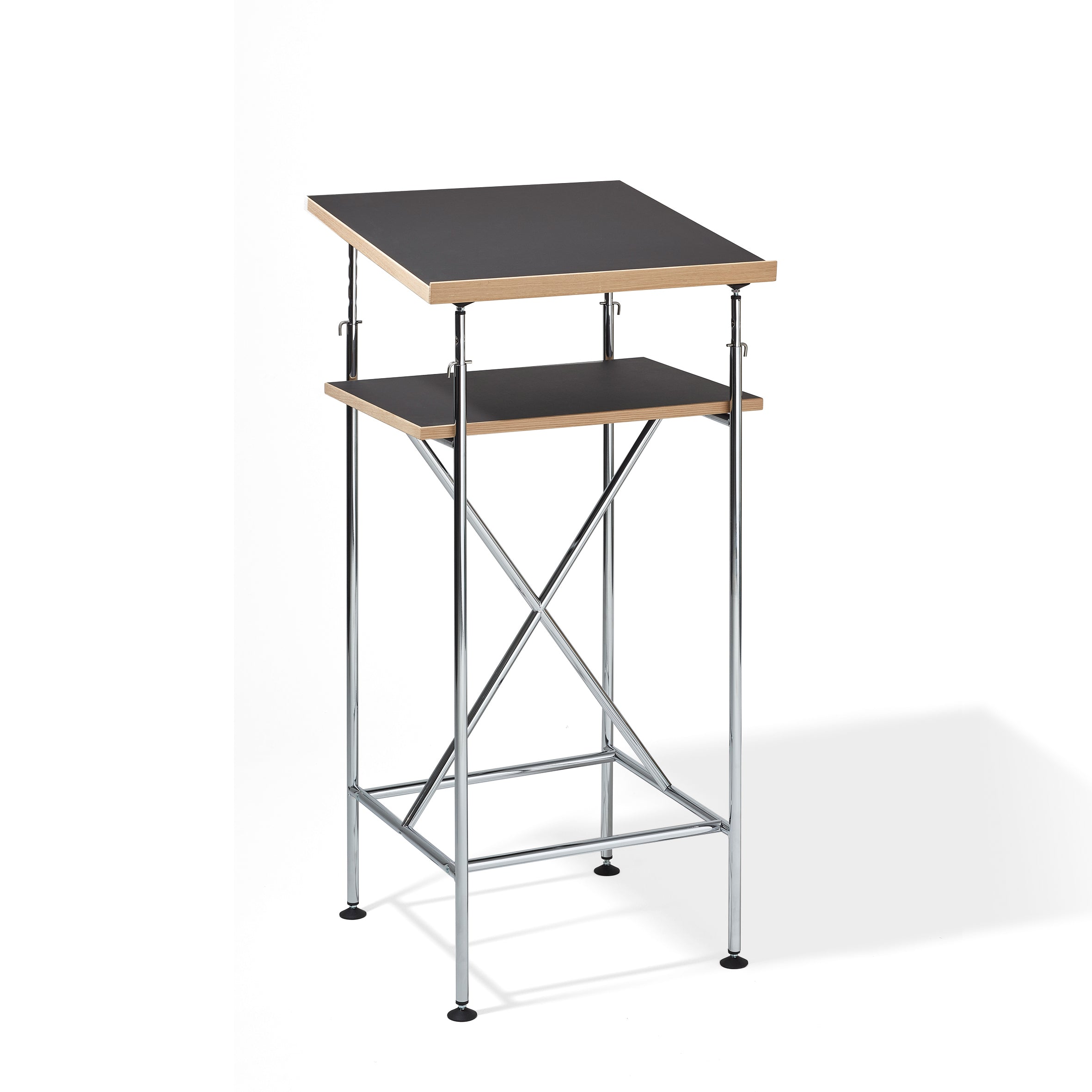 The Milla High Desk by Richard Lampert is a modern black podium resembling an Eiermann table. It features a slanted top, elevated shelf, and X-shaped metal supports for active sitting and standing, all in a minimalist design.