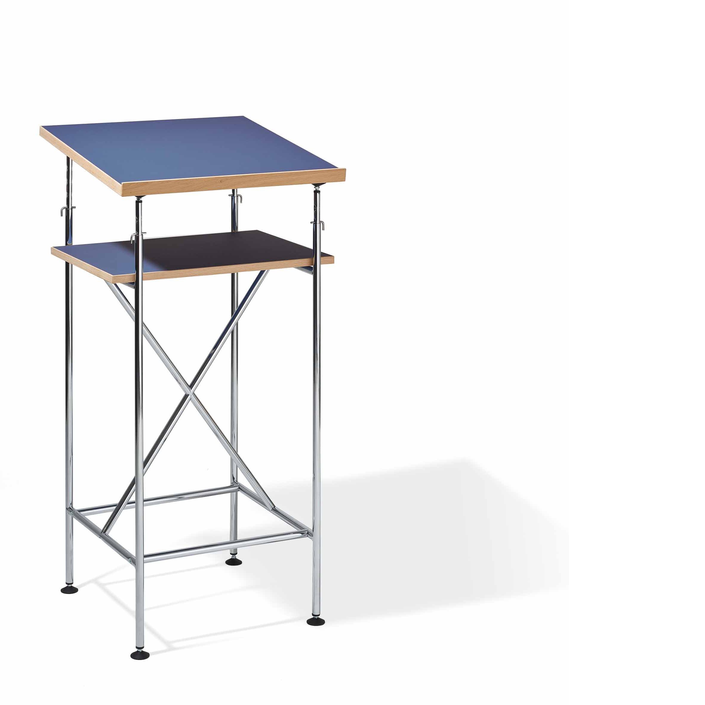 The Milla High Desk by Richard Lampert features a design similar to an Eiermann table with a modern, minimalistic style. It has a blue top, metal frame, wooden accents, and dual-tier construction with a smaller black shelf underneath, all set against a plain white backdrop.