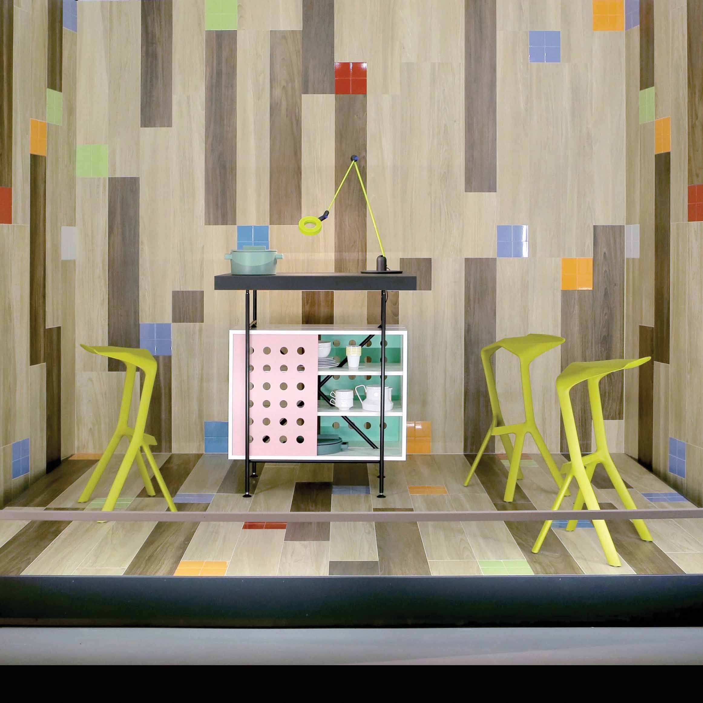 A modern dining area features multicolored tiled walls and floor. The Milla Bistro Table by Richard Lampert, with its geometric design, holds a blue bowl and a yellow lamp. Four uniquely shaped green stools surround the table, adding to the eclectic atmosphere with smart storage solutions.