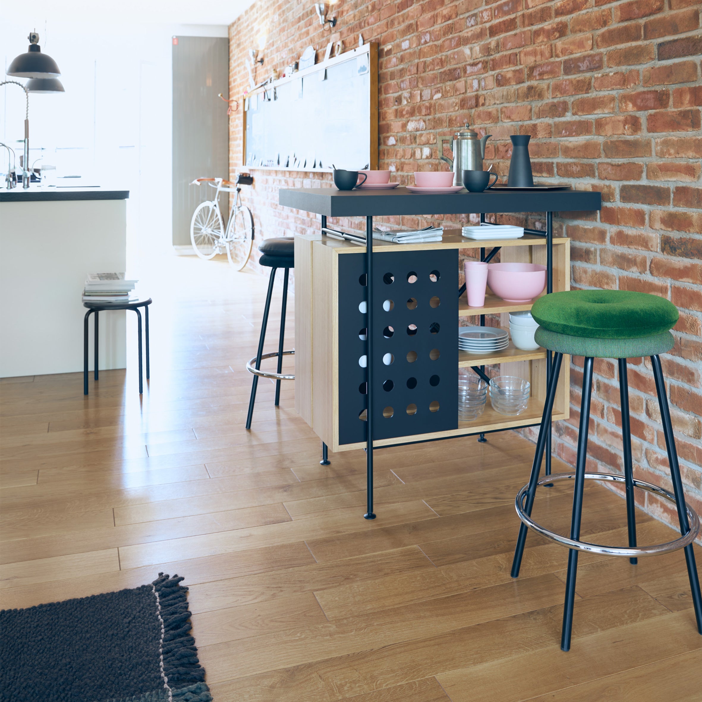 The modern kitchen boasts a wooden floor and brick wall. A Richard Lampert Milla Bistro Table doubles as a bar featuring kitchenware and a black metal frame. Two barstools, one with a green cushion, flank it. A bicycle and ample storage complete the background scene.