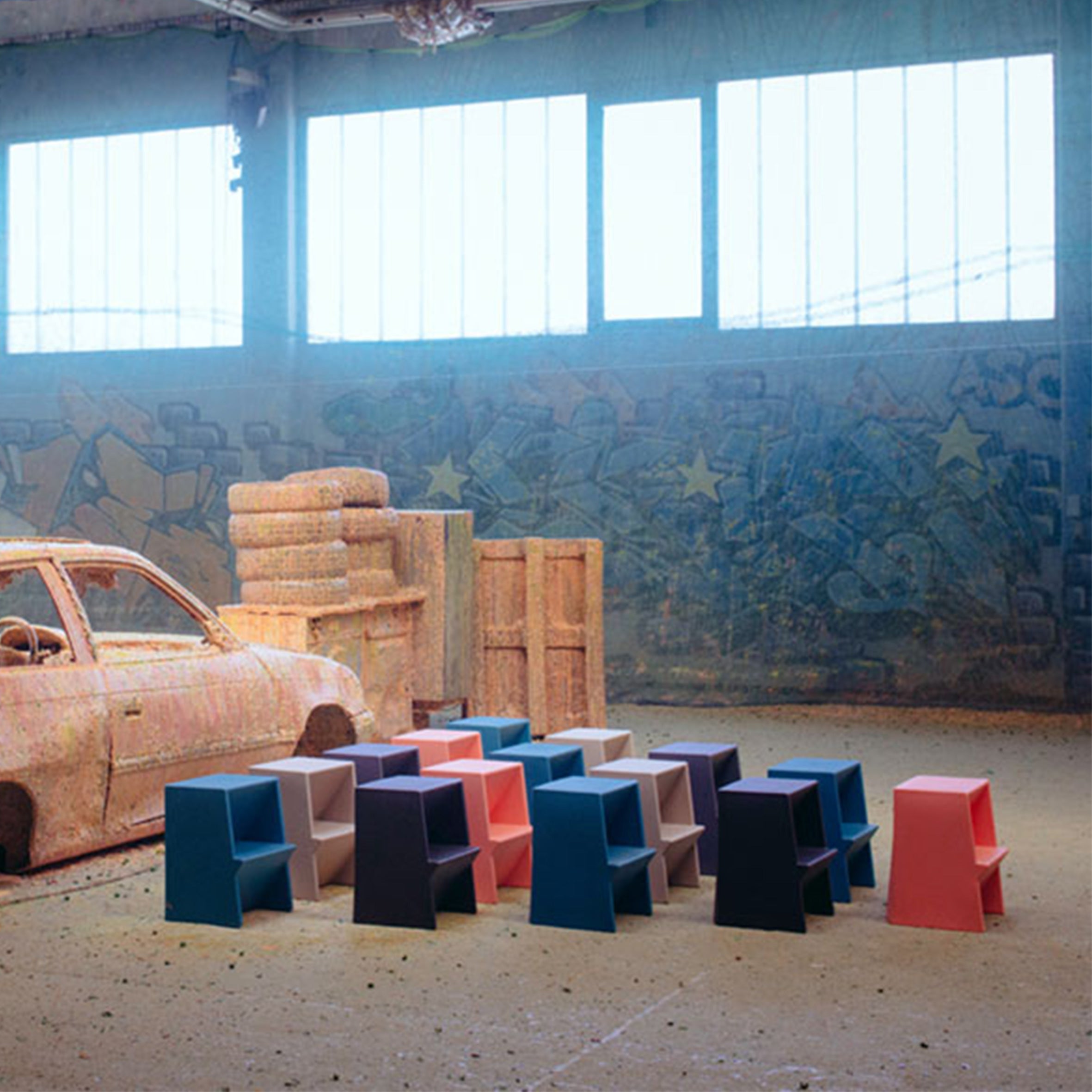 Colorful Richard Lampert Mono Stepstools, crafted from recyclable polyethylene, are stacked at the center of an industrial-style room with large windows and graffiti. An old car and stacked pillars on the left enhance the urban artistic vibe.