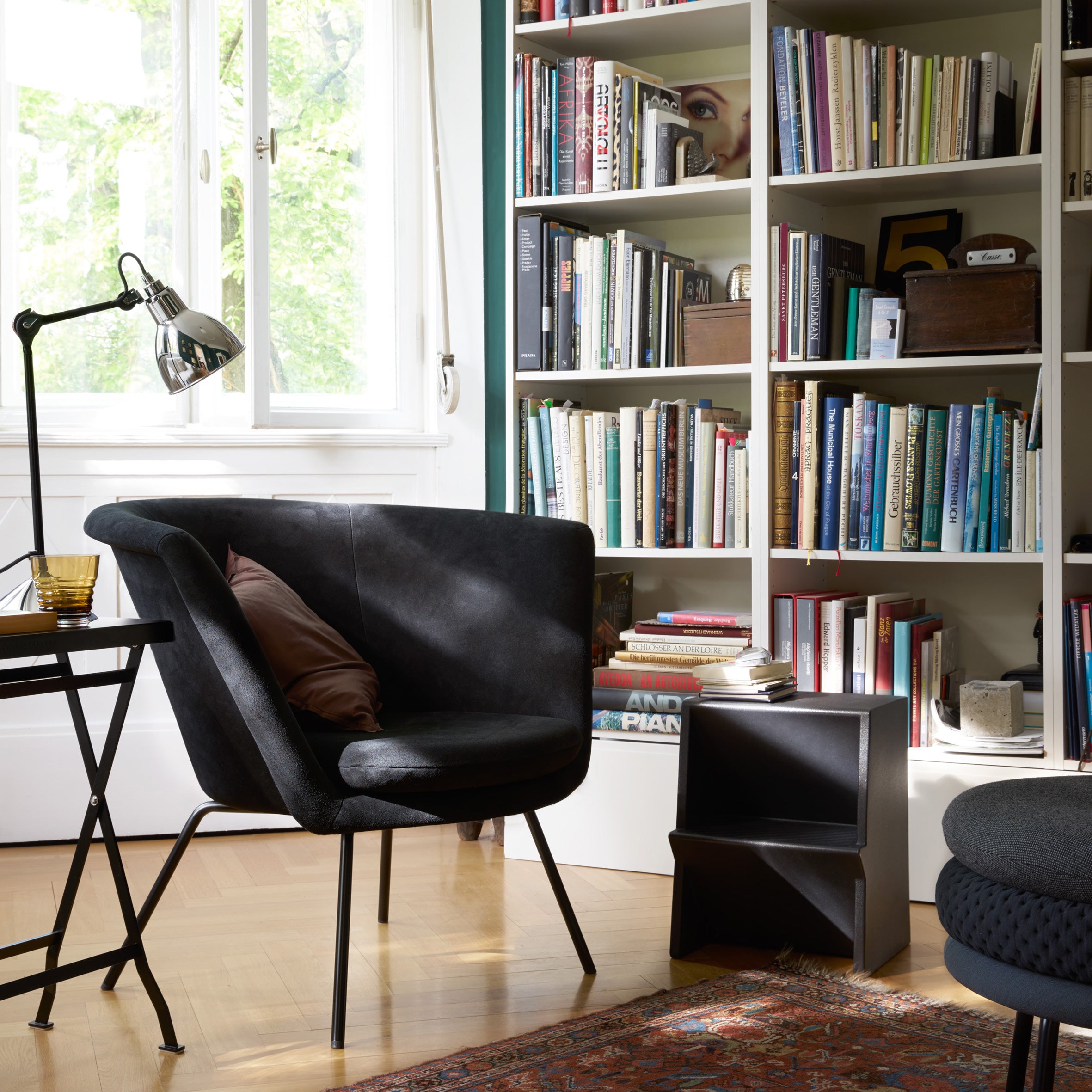 A cozy reading nook featuring a black armchair by a tall white bookshelf, complemented by Richard Lamperts Mono Stepstool for extra seating. A small folding table holds a lamp and glass, as natural light streams through the window onto the patterned rug on the wooden floor.