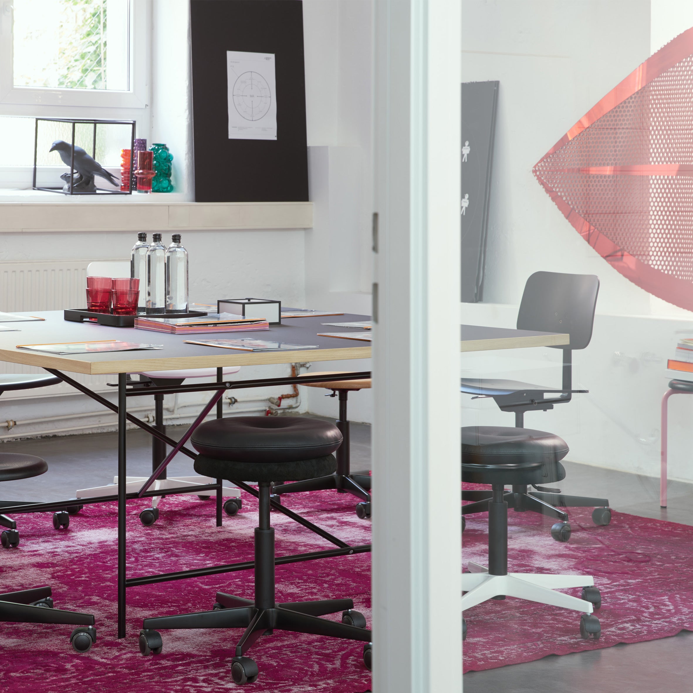 A modern office space showcases a large table with Mr. Round Swivel Stools by Richard Lampert, all set atop a magenta rug. The table is decorated with red glasses, a notebook, and water bottles. Artworks adorn the walls, while a geometric red sculpture enhances the corners appeal.