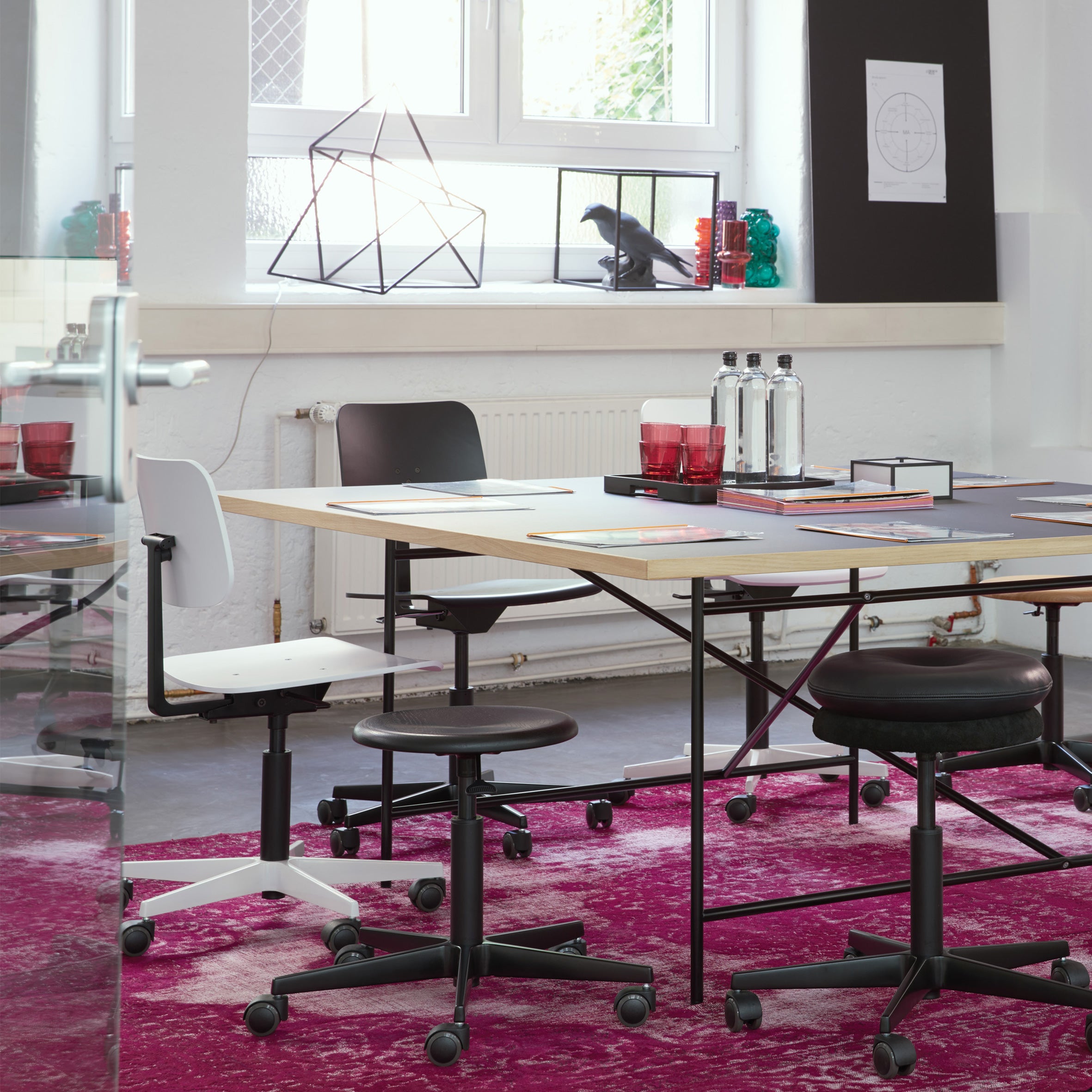 In a modern conference room, a large table on a vibrant pink rug is surrounded by Richard Lamperts Mr. Square Task Chairs and designer stools for superior comfort. The table is set with notebooks, glasses, and carafes, while a window showcases geometric decor and lush plants in the background.