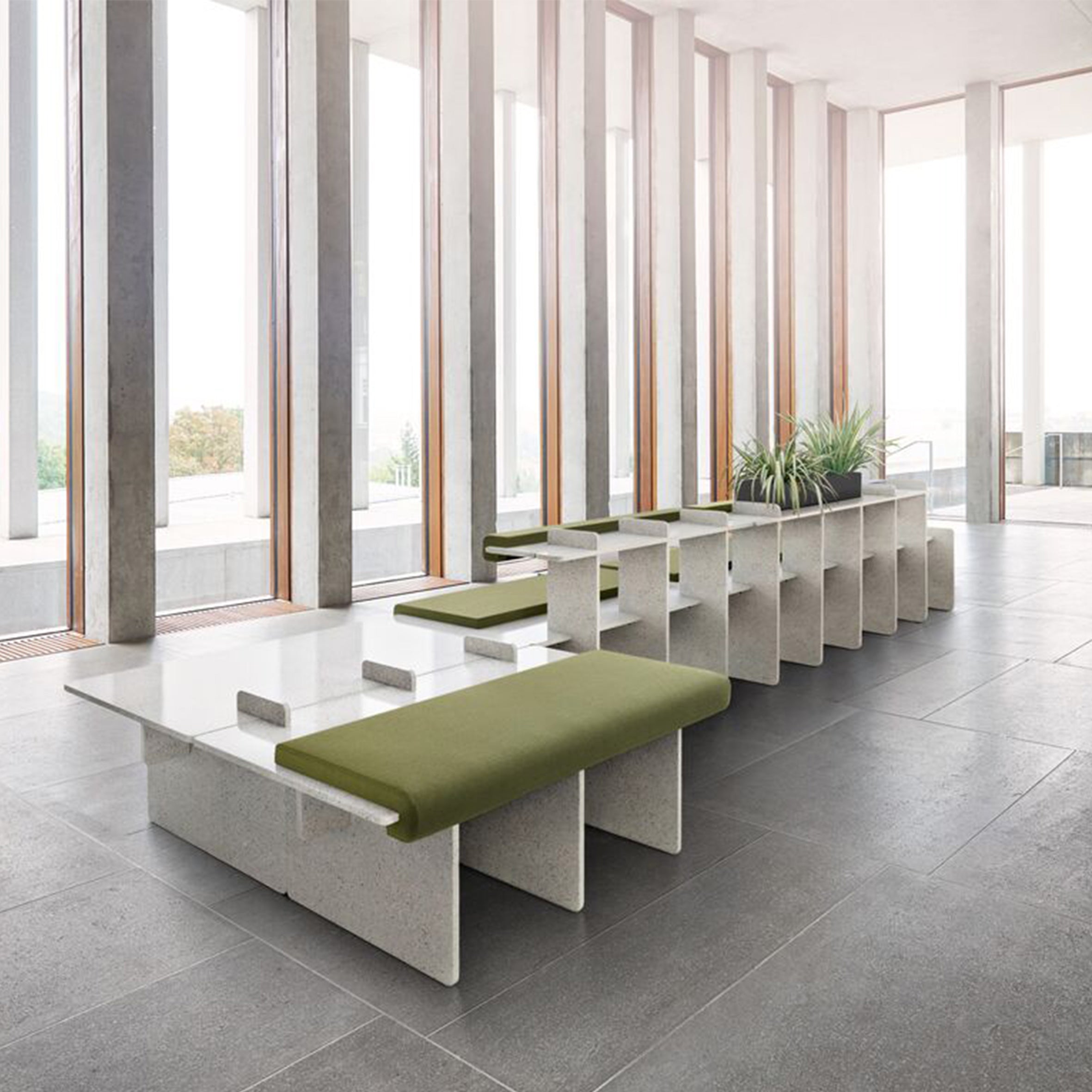 The modern seating area features Richard Lamperts Pip furniture with modular two-tone benches and green cushions, complemented by sleek gray stone-like tables. Tall windows brighten the space with plants on tables against a backdrop of light-colored pillars and walls.