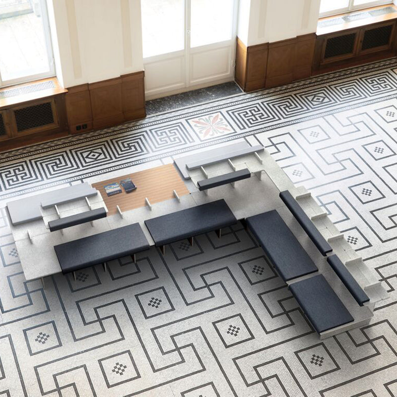 Richard Lamperts Pip offers a fireproof, modular L-shaped seating setup in a spacious room with geometric-patterned floor tiles. It includes black and gray tables and cushions. Large windows framed with wooden paneling are featured in the background.