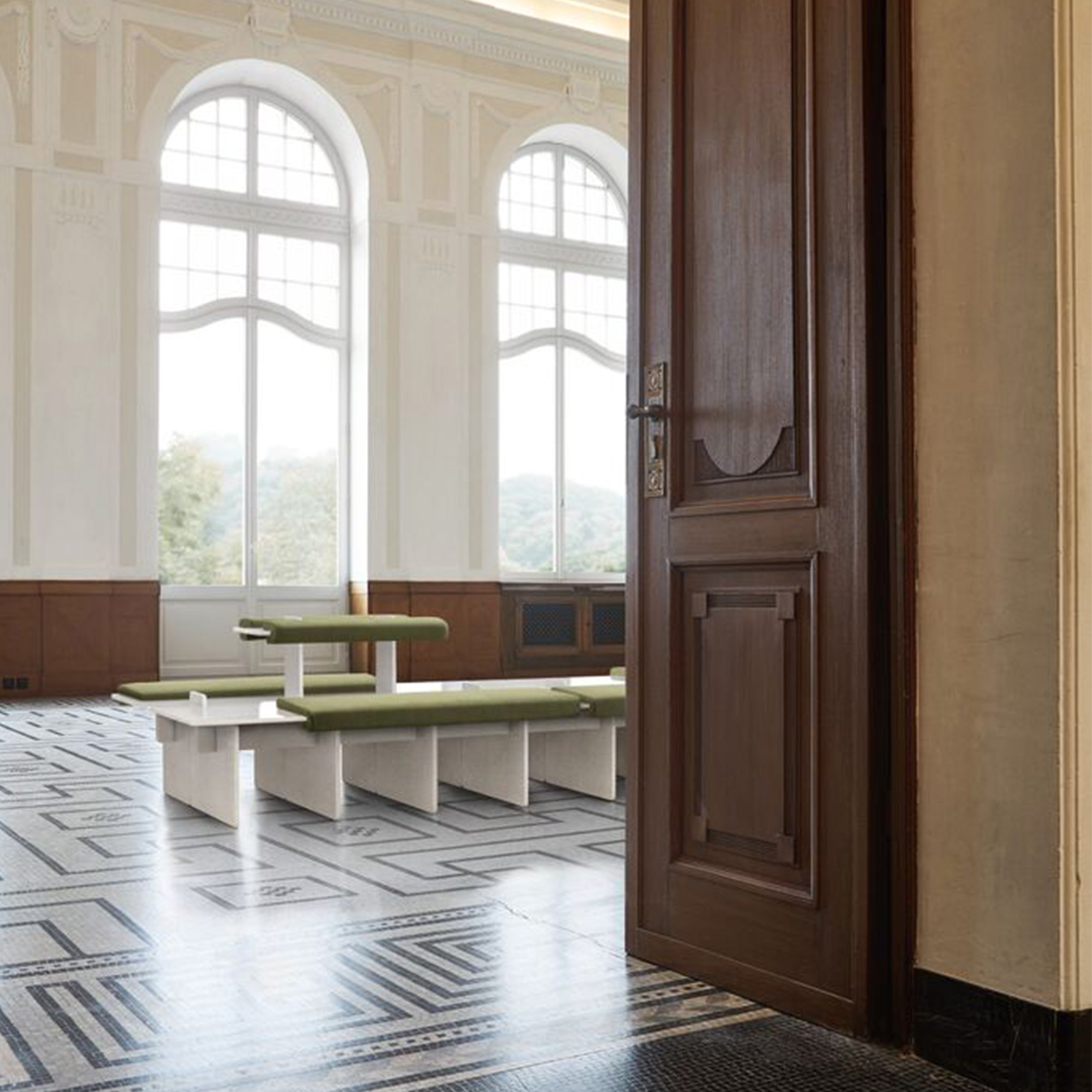 Beyond the open wooden door lies a bright room with large arched windows. The space features the green, variable-seating Pip by Richard Lampert and a floor adorned with black-and-white geometric tiles. Trees are visible through the windows.