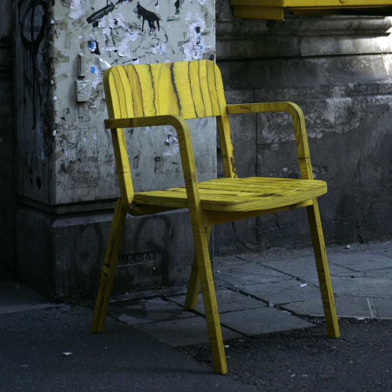 The Prater Chair by Richard Lampert, with its vibrant yellow birch plywood crafted through CNC milling, stands on a dimly lit, weathered sidewalk beside torn posters and graffiti, creating a striking contrast between urban decay and bright color.