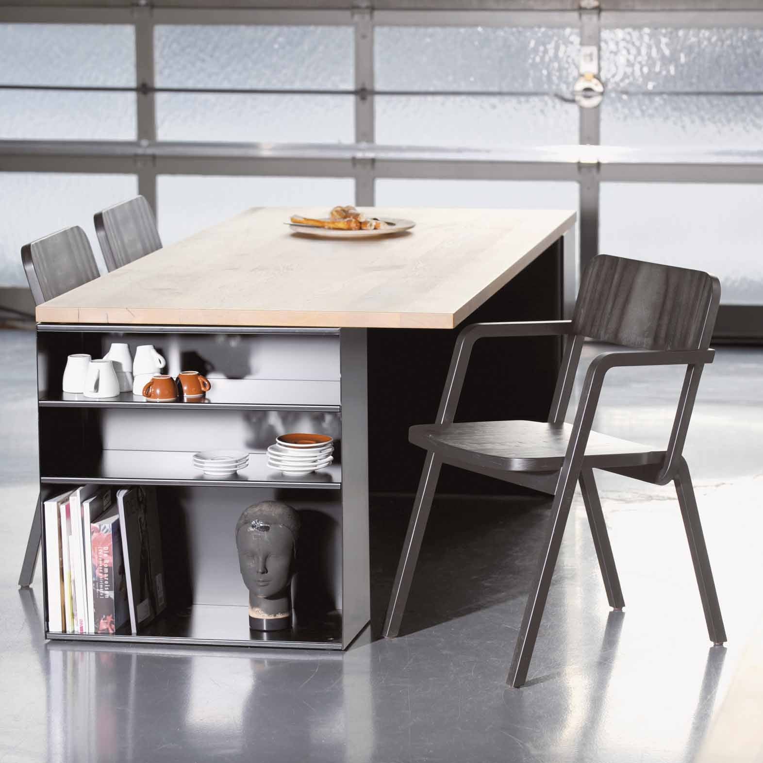 A modern dining setup features a sleek wooden top complemented by stylish black and Prater chairs by Richard Lampert. Beneath, shelves display dishes, a black bust, and books. A plate with baked goods rests atop the table in a room adorned with glossy flooring and an elegant frosted glass wall.