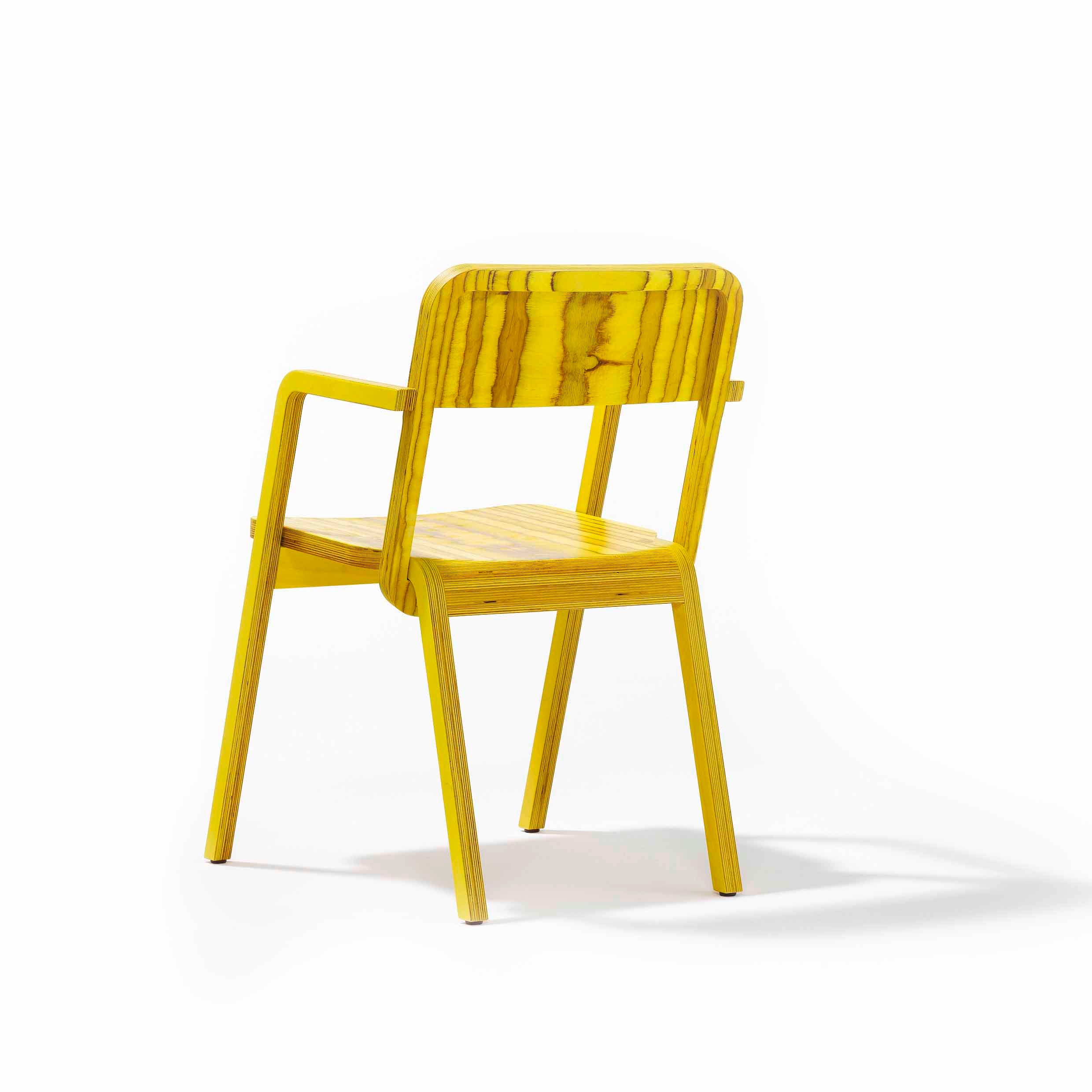 The Prater Chair by Richard Lampert is a yellow birch plywood piece with a minimalist design, featuring straight legs and a slatted backrest. It casts a light shadow on the white background, reminiscent of Viennese wooden chairs, and is viewed from the back at an angle.