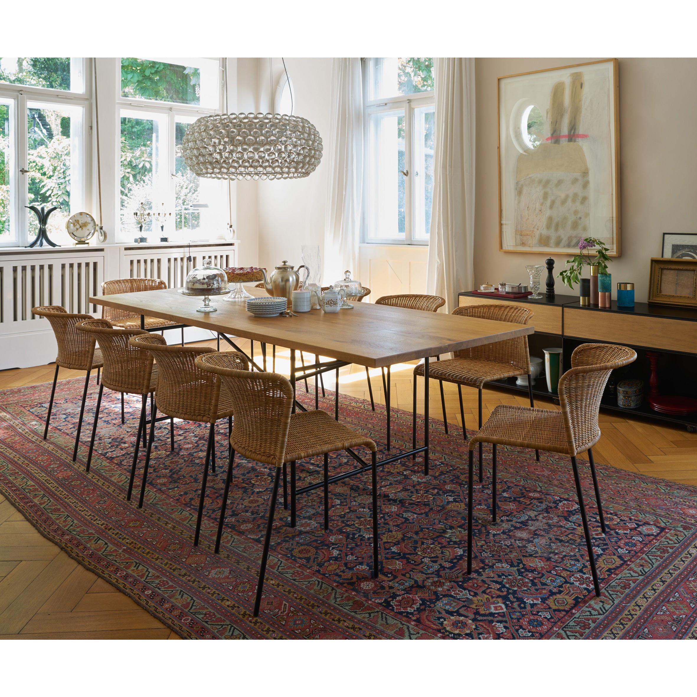 A dining room showcases the Santa Lucia wooden table by Richard Lampert, surrounded by eight tubular steel chairs on a patterned red and blue rug. A modern circular chandelier hangs above, while large windows with white curtains illuminate the artwork and a decor-adorned sideboard.
