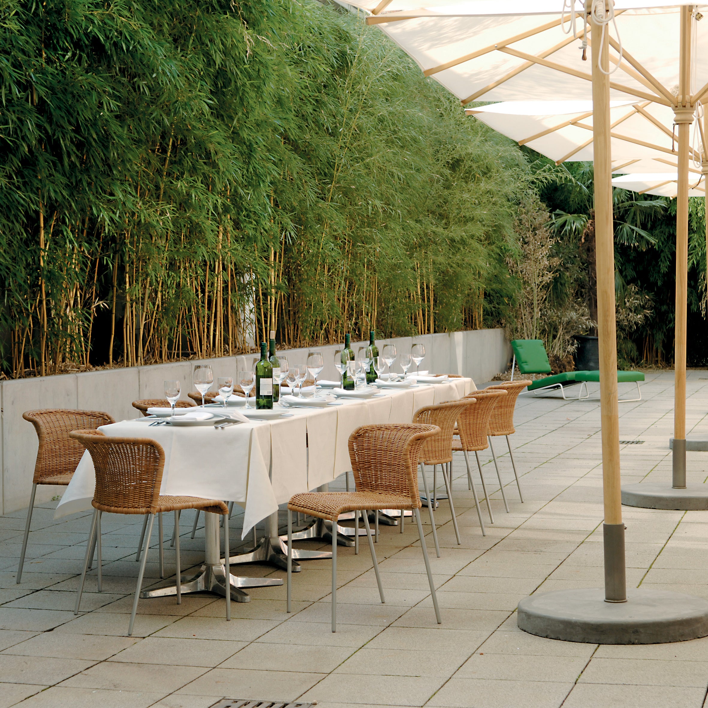 An outdoor dining space features the Santa Lucia table by Richard Lampert, set with wine glasses, bottles, and white tablecloths. It is surrounded by rattan chairs and green plants under large umbrellas on a tiled patio.