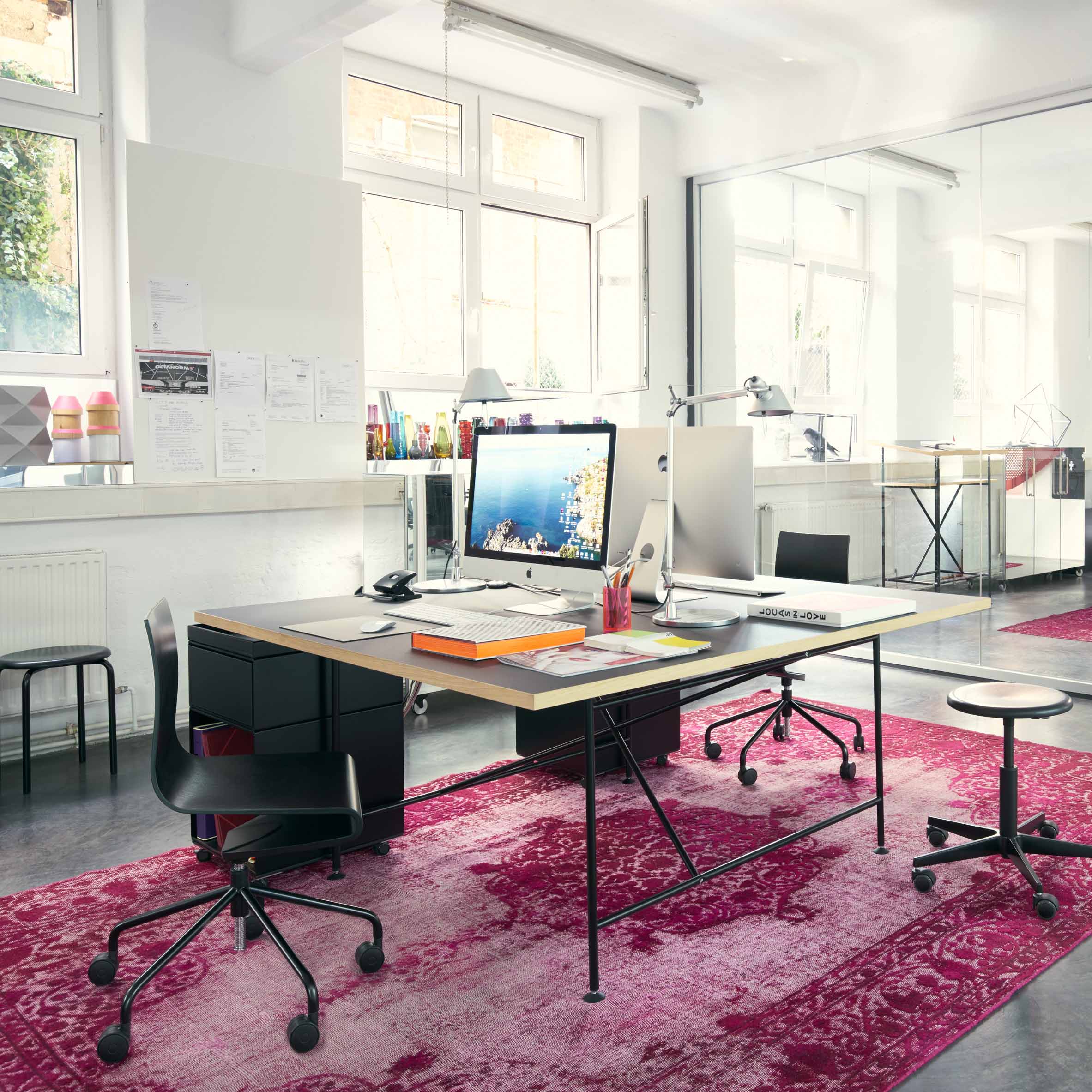 A modern office space features two desks, each with a Seesaw Task Chair by Richard Lampert. Red patterned rugs cover the floor, and large windows let in natural light. Bright, minimalist furnishings enhance the inviting atmosphere.