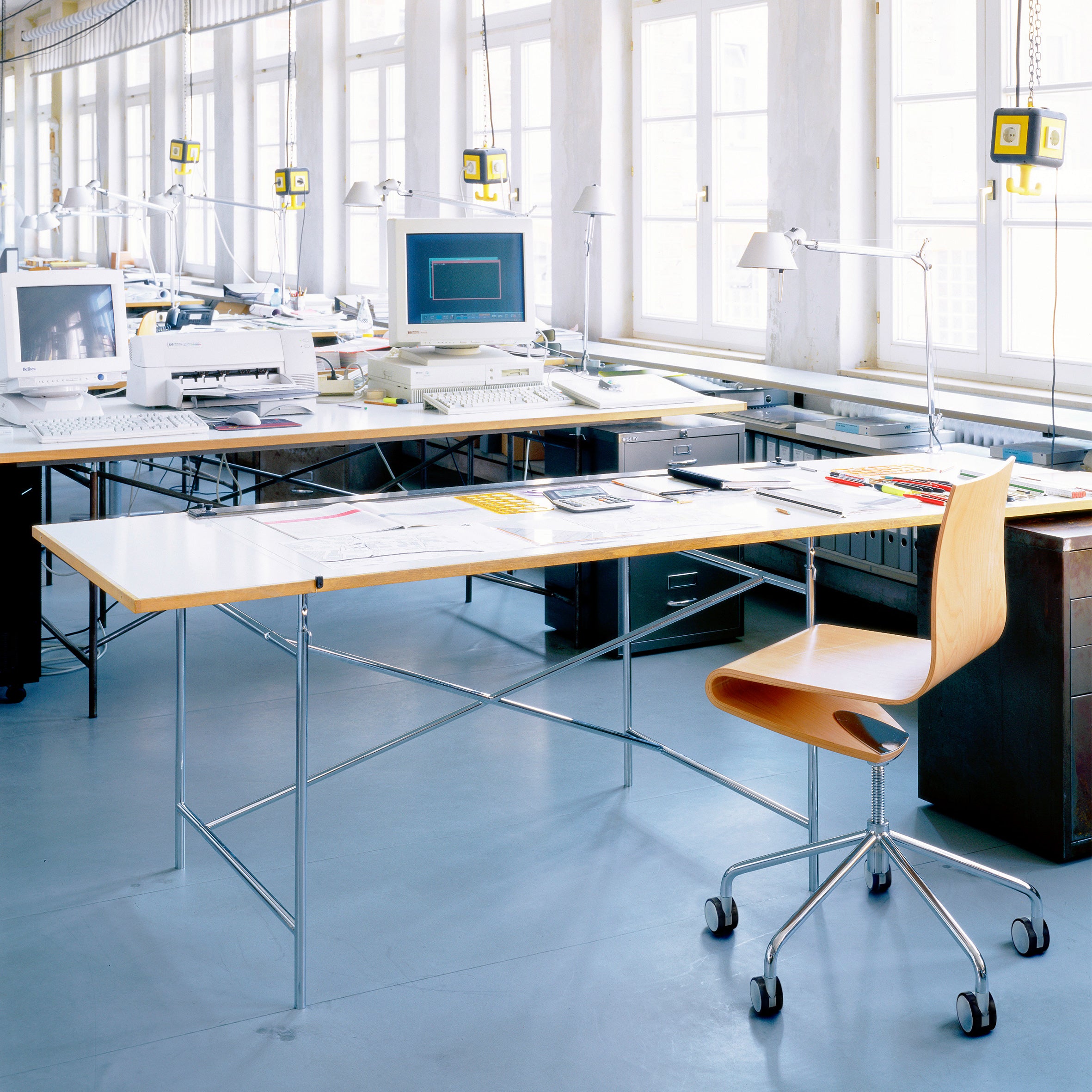 A spacious, brightly lit office with large windows features a modern, minimalistic setup including the ergonomic Seesaw Task Chair by Richard Lampert, sleek desks, scattered papers, desktop computers, a printer, lamps, and measuring tools hanging from the ceiling.