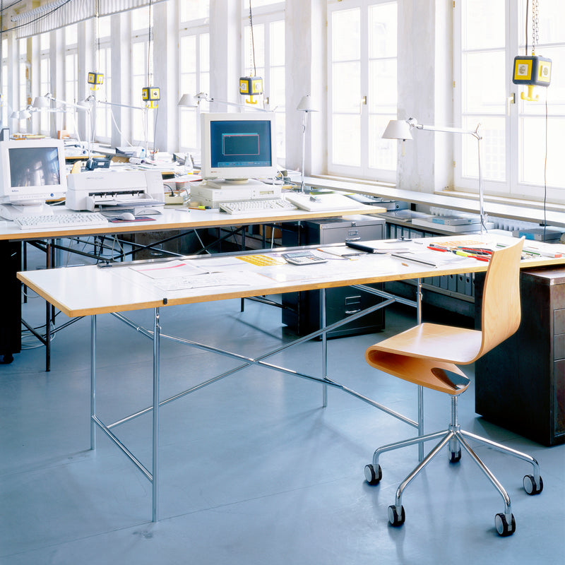 A spacious, brightly lit office with large windows features a modern, minimalistic setup including the ergonomic Seesaw Task Chair by Richard Lampert, sleek desks, scattered papers, desktop computers, a printer, lamps, and measuring tools hanging from the ceiling.