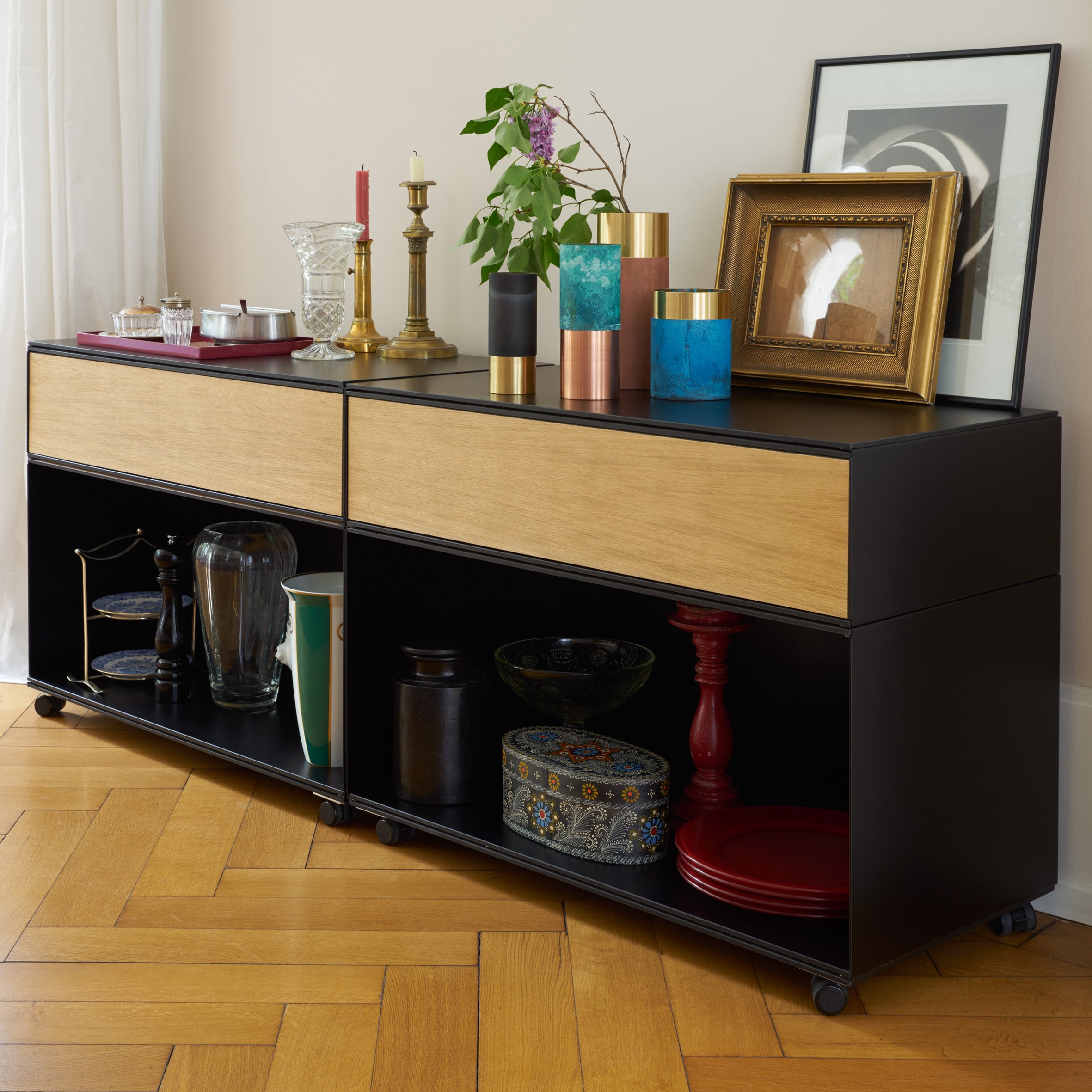 The Stak Trolley System by Richard Lampert, featuring black and wood design, stylishly displays eclectic decor such as colorful vases, candles, framed art, and red plates on a wooden floor. Solid oak shelves ensure functionality with storage innovation while a plant enhances its modern appeal.