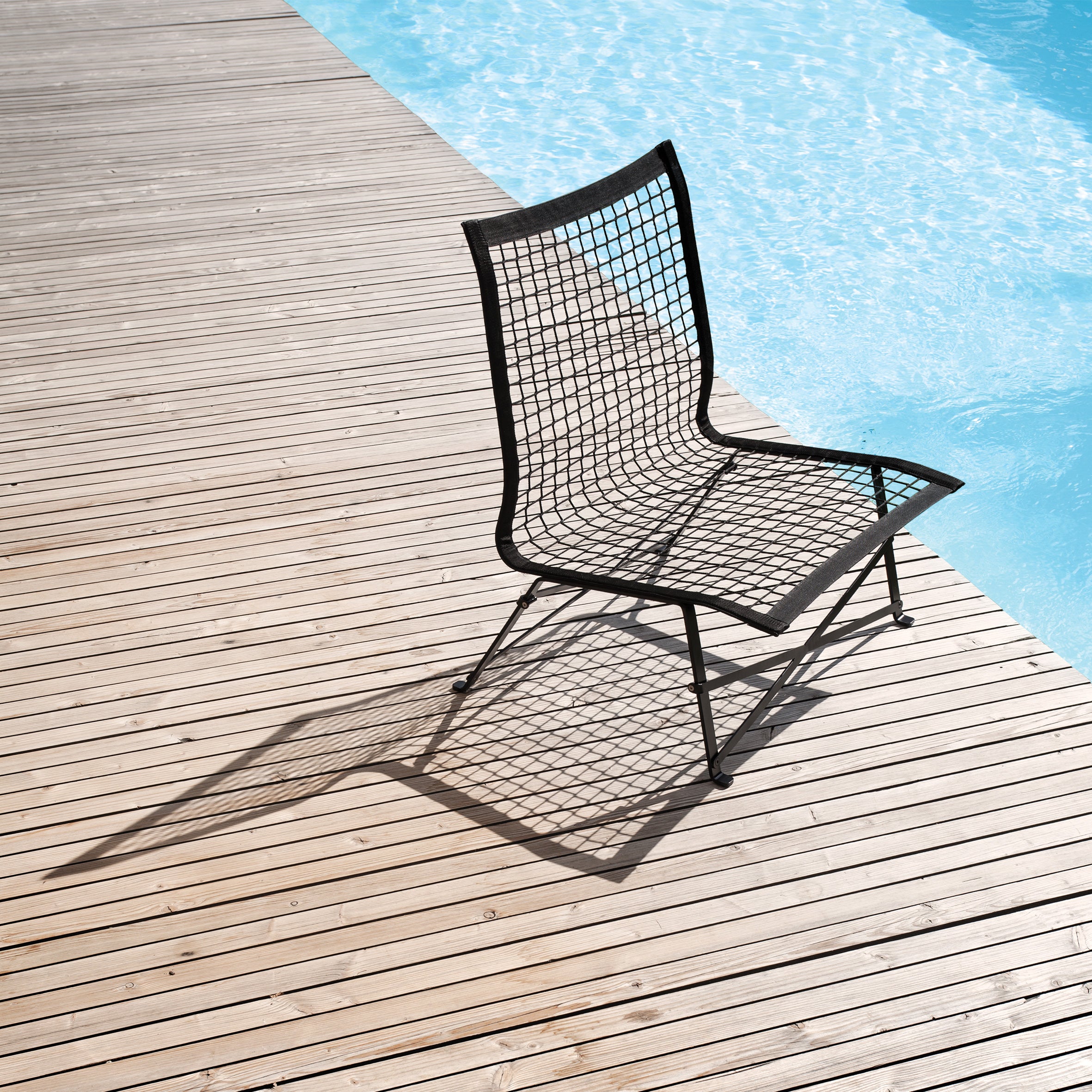 The Tie-Break Lounge Chair from Richard Lampert, featuring a tall backrest and black metal mesh design, casts a shadow on the wooden deck beside a clear blue pool under bright sunlight, epitomizing elegant garden furniture.