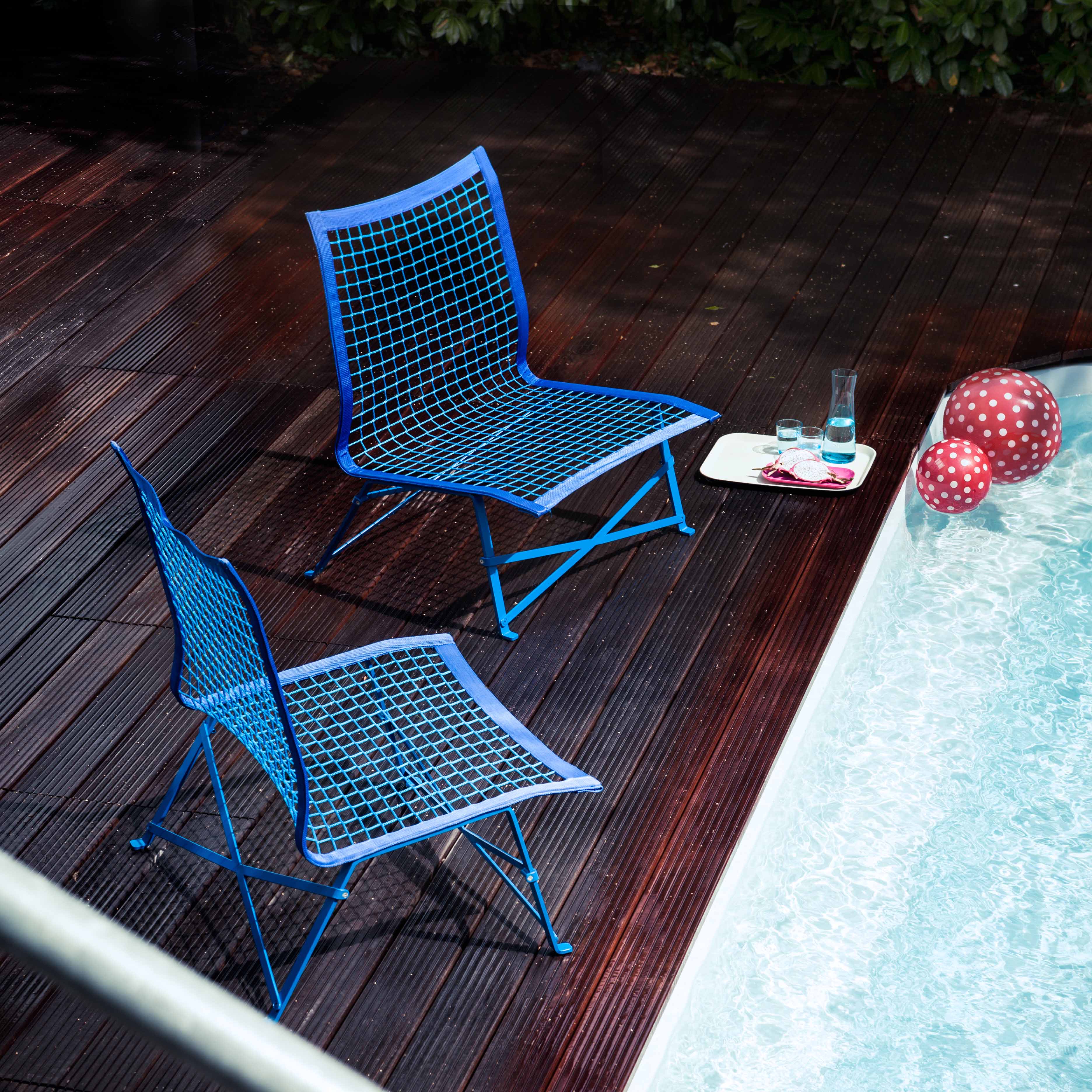 Two Tie-Break Lounge Chairs by Richard Lampert, in blue mesh, are on a wooden deck beside the pool. A tray with a drink and glass sits nearby as red and white polka dot inflatable balls float in the water. Lush greenery enhances this serene garden furniture setting for relaxation.