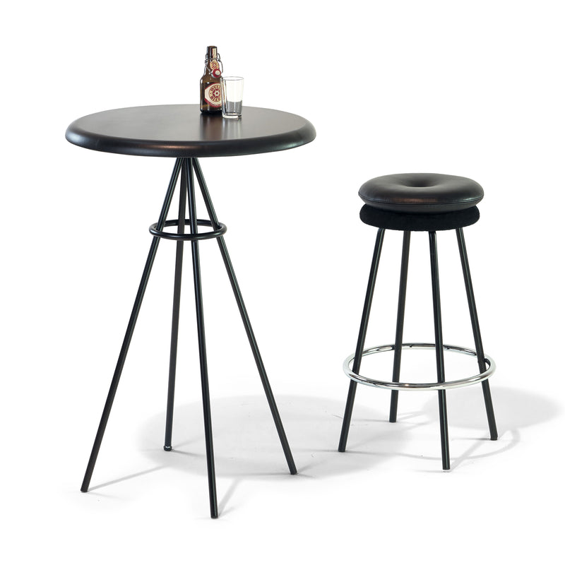 The Richard Lampert Tom Bar Table, featuring a black top and slender metal legs with a contrasting solid oak surface, is paired with a matching bar stool that has a cushioned seat. The table holds a small glass and bottle.