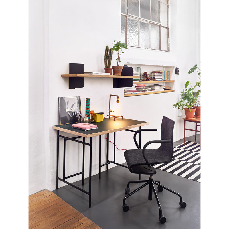 A minimalist workspace showcases a sleek Richard Lampert Seesaw Task Chair, a desk with a lamp, notebooks, and a record. Shelves above display books, plants, and decor. A striped rug adorns the floor as sunlight pours through the window in the background.
