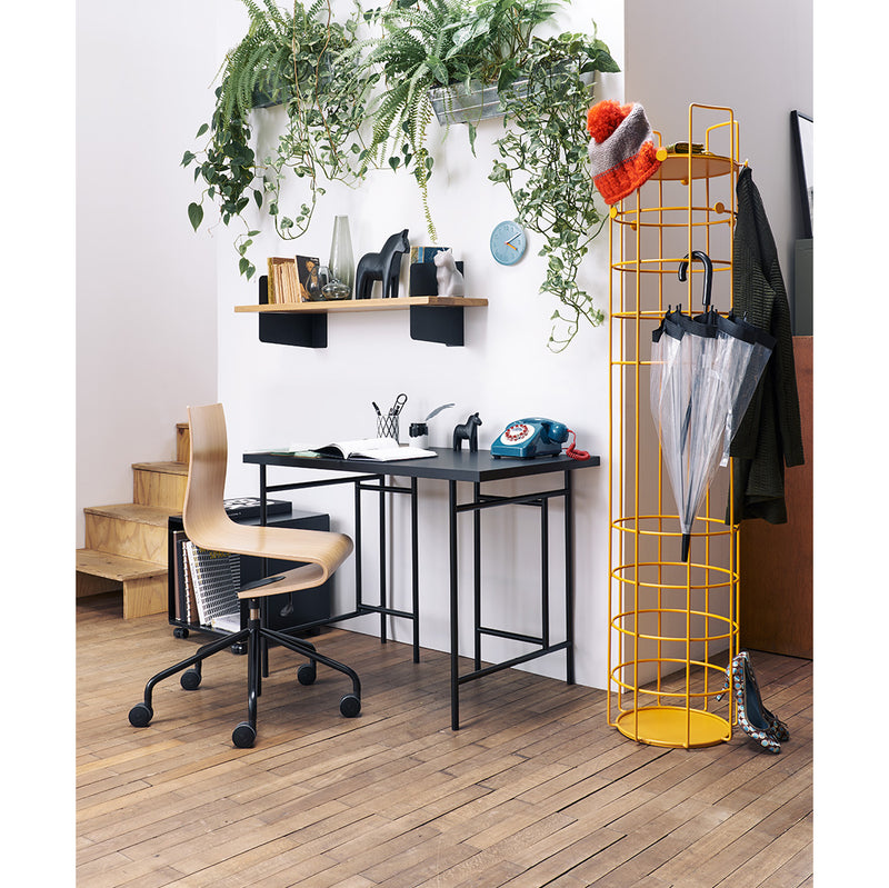 The cozy home office features the Richard Lampert Atelier sleek black desk and chair, complemented by potted plants and books on wooden shelves. Steel brackets add elegance, while a vibrant yellow coat rack holds essentials. The wooden floor enhances warmth in this minimalistic space.