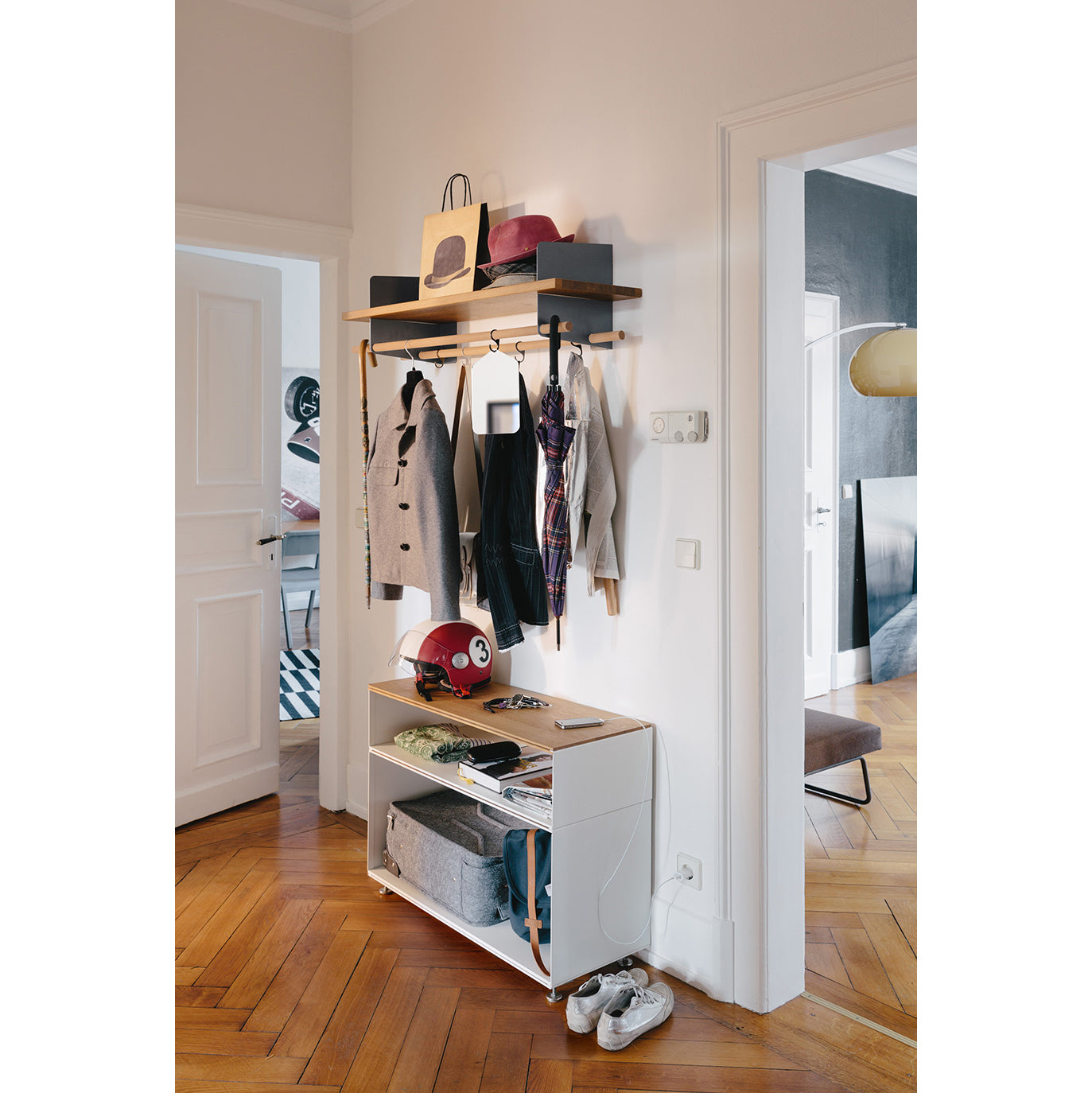 A stylish hallway with a wooden floor showcases innovative storage, featuring Richard Lamperts Stak Trolley System. Wall-mounted solid oak shelves and coat racks hold jackets, bags, and scarves on hooks, a white cabinet stores hats and baskets below, while shoes rest on the floor. An open door reveals another room.