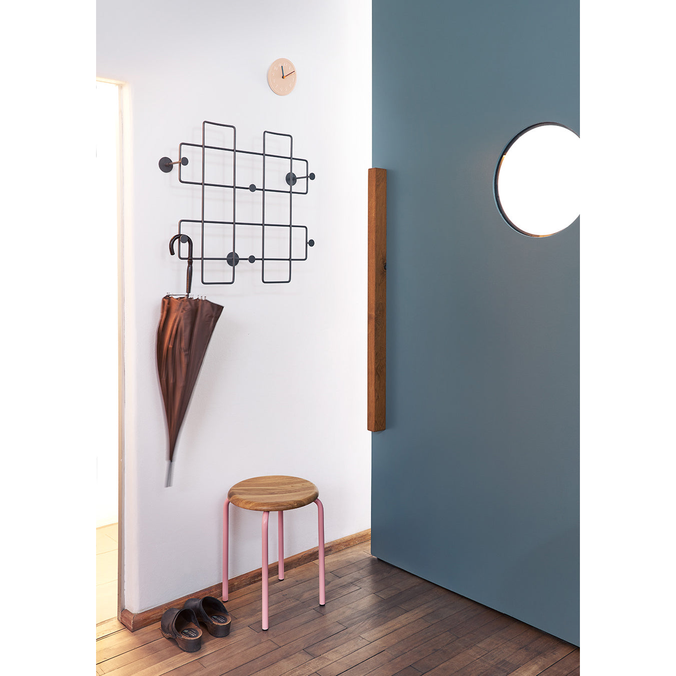A minimalist hallway showcases a wooden floor, a Richard Lampert Tom Stackable Stool with a pink frame, shoes, and an umbrella on a geometric wall rack. A partly open blue door with a round window and a small wall clock are also visible.