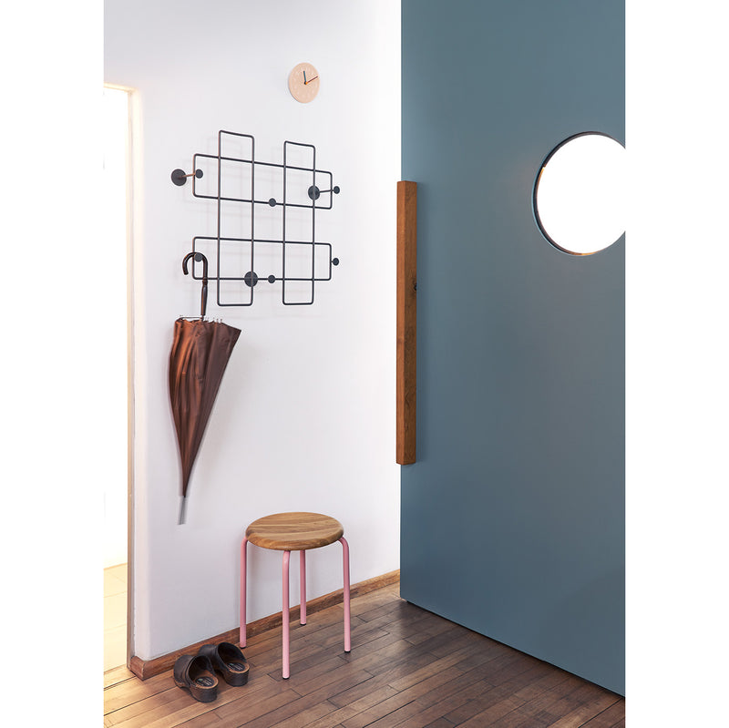 A minimalist hallway showcases a wooden floor, a Richard Lampert Tom Stackable Stool with a pink frame, shoes, and an umbrella on a geometric wall rack. A partly open blue door with a round window and a small wall clock are also visible.