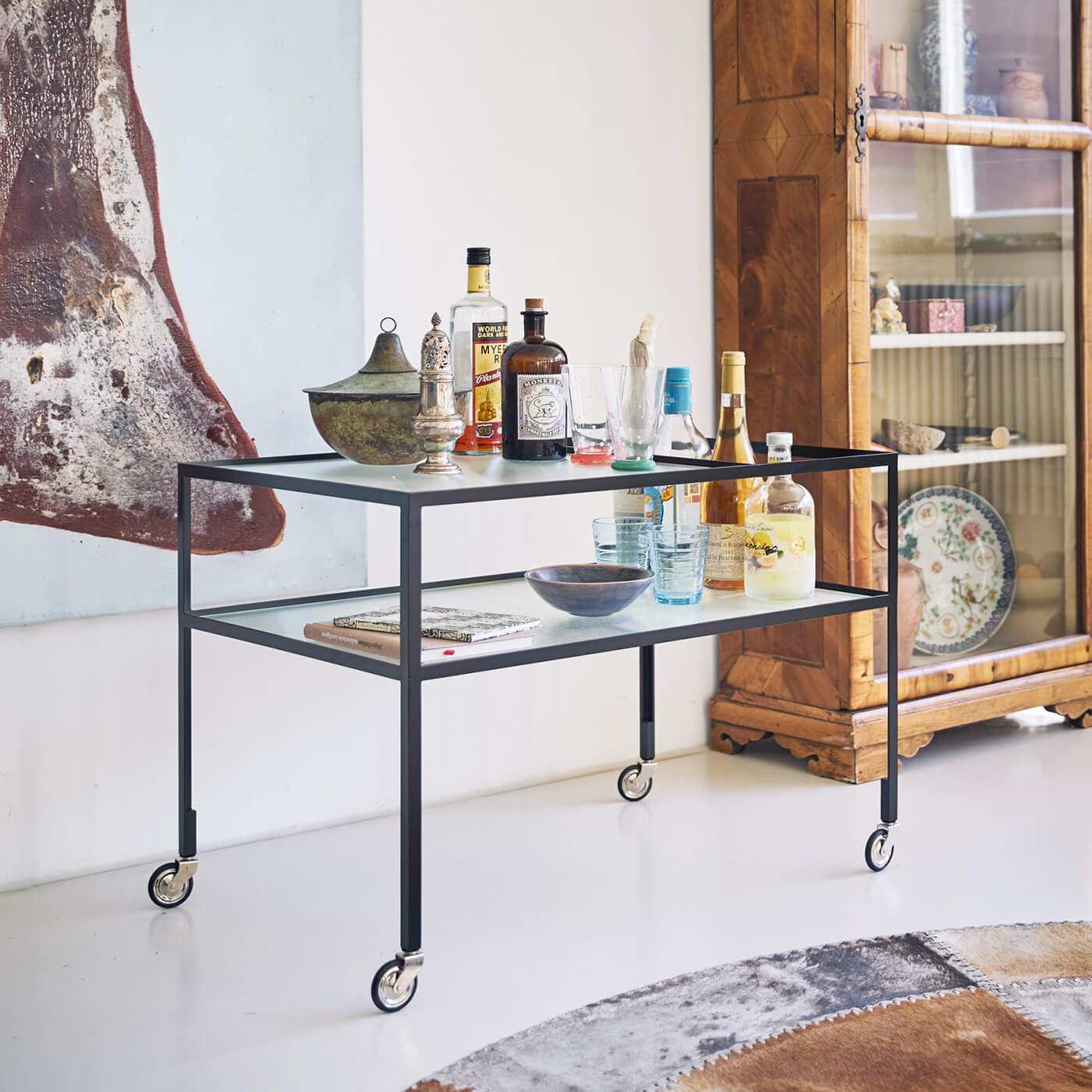 The Richard Lampert Bar Trolley is a sleek black metal cart featuring a minimal steel design and wheels, perfect for storing liquor bottles, glasses, and decor. Its situated beside a wooden cabinet with glass doors, enhanced by a textured abstract painting on the white wall behind it.