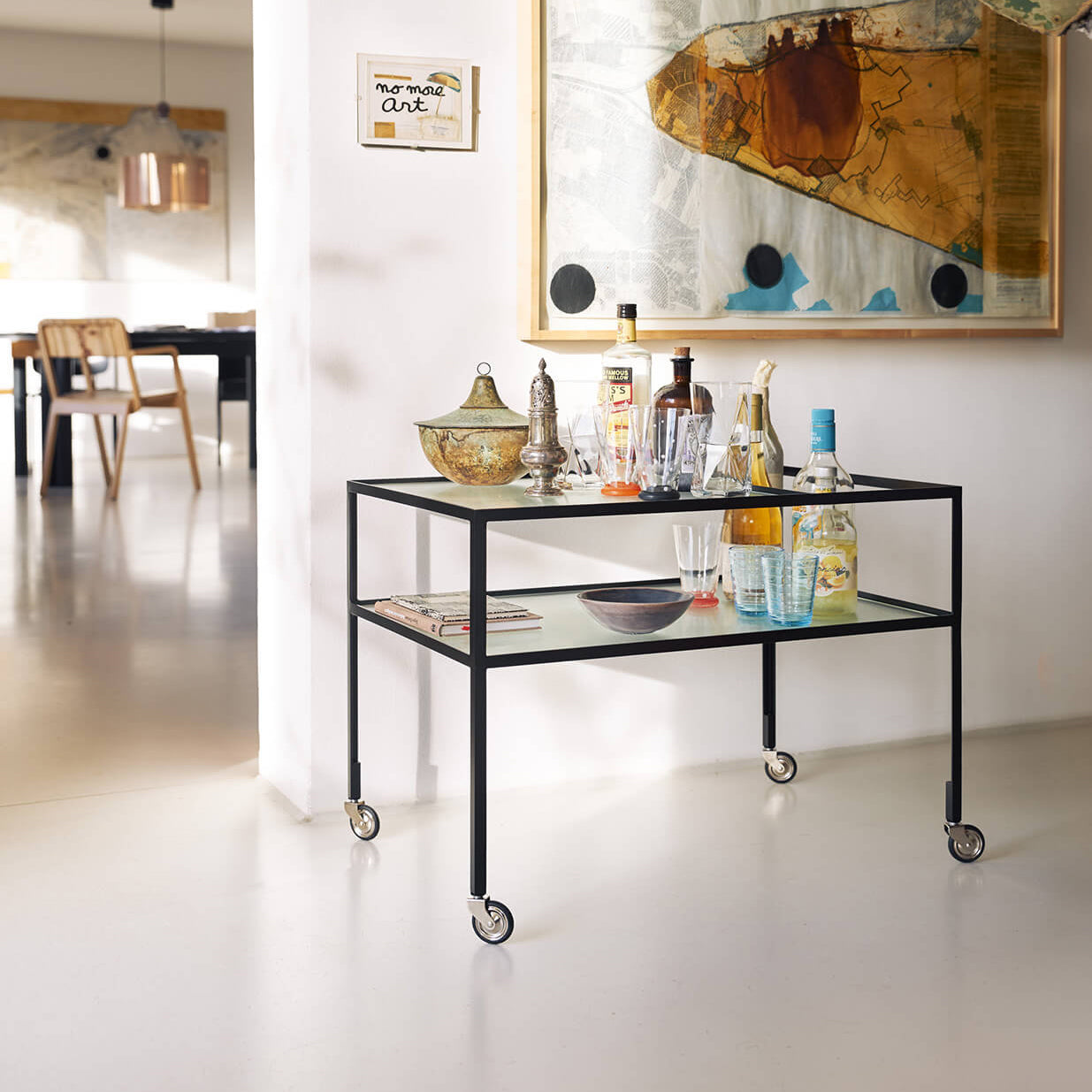 A Richard Lampert Bar Trolley with various bottles and glasses is set against a wall featuring abstract artwork. Its sleek, minimal black steel design adds elegance to the modern room, which includes minimalistic furniture like a wooden chair and dining table in the background.