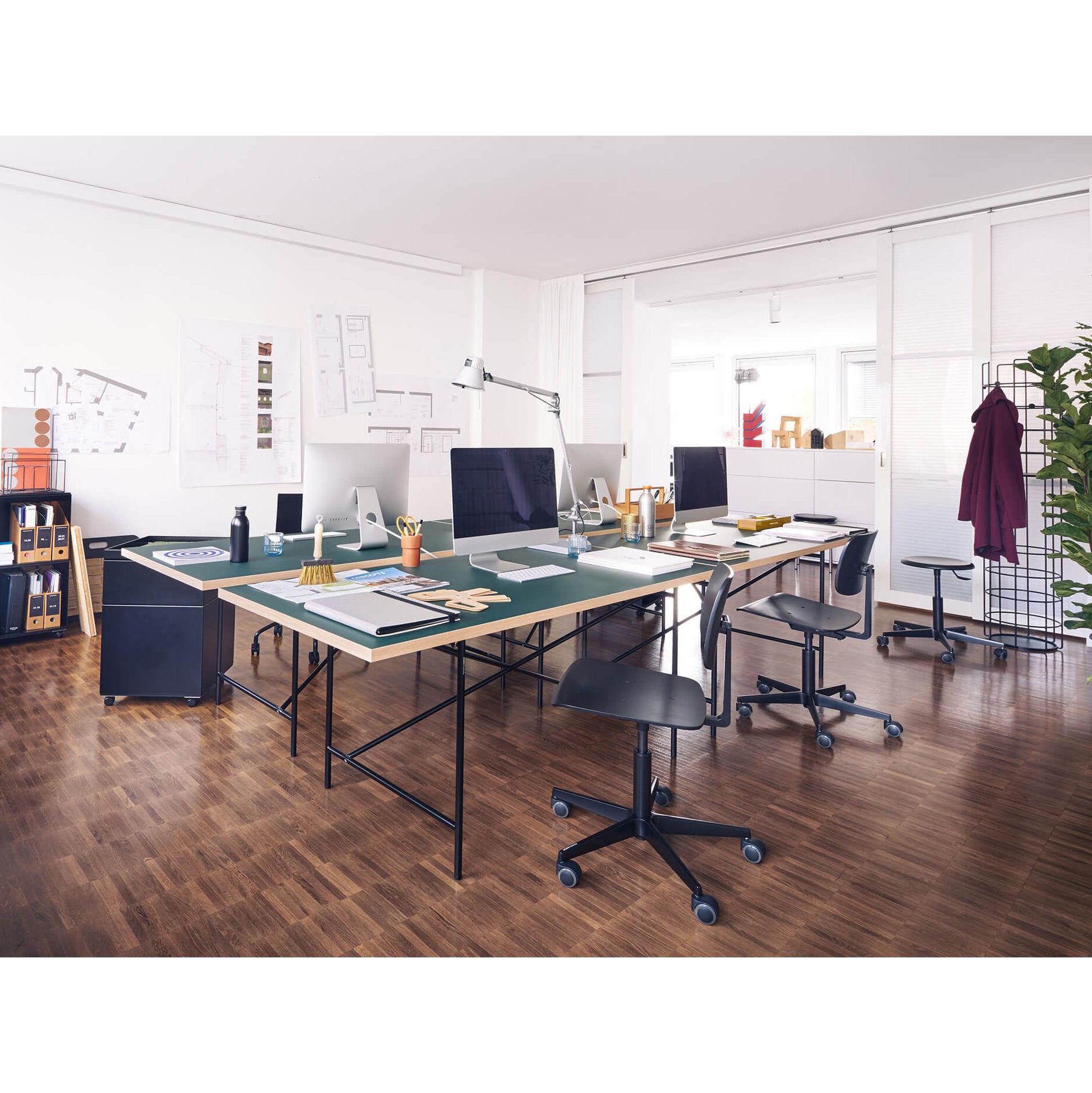 A spacious, modern office features large desks, height-adjustable computers, design tools on a wooden floor, and Mr. Square Task Chairs by Richard Lampert. Architectural plans decorate the walls, with a coat rack and potted plant in the corner. The room is bathed in bright natural light.