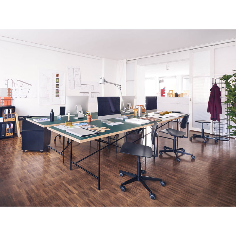 A spacious, modern office features large desks, height-adjustable computers, design tools on a wooden floor, and Mr. Square Task Chairs by Richard Lampert. Architectural plans decorate the walls, with a coat rack and potted plant in the corner. The room is bathed in bright natural light.