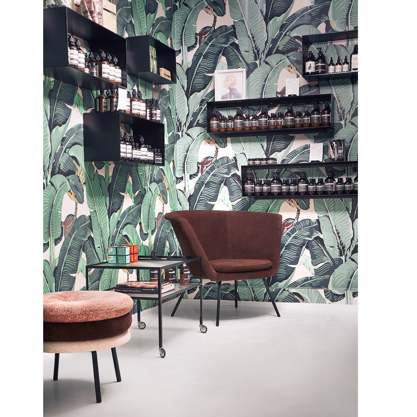 The room showcases tropical leaf wallpaper and sleek black steel shelves displaying bottles. A brown upholstered chair accompanies a small book-adorned table, exuding timelessness. Nearby, Richard Lamperts Bar Trolley graces the light floor, embodying Herbert Hirche’s aesthetic.